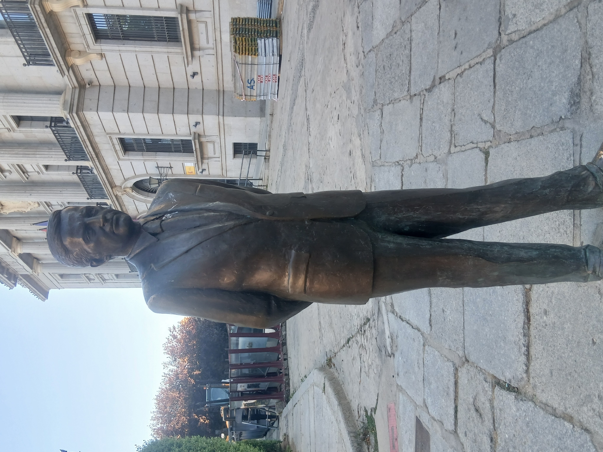 A full-body bronze statue of a man in a suit stands on a paved walkway in front of a stone building. The man is facing to the right of the frame with his hands clasped behind his back. The building behind him has a classical facade with columns, balconies, and barred windows. In the middle ground to the right of the building, a stack of scaffolding is visible. To the left of the statue, a sidewalk and a patch of grass are present, with a large construction dumpster and some portable barriers further back. The ground is paved with large, irregular stone slabs. The sky is clear and blue. This statue is located in the Plaza de Santa Ana in the city of Ávila, Spain.