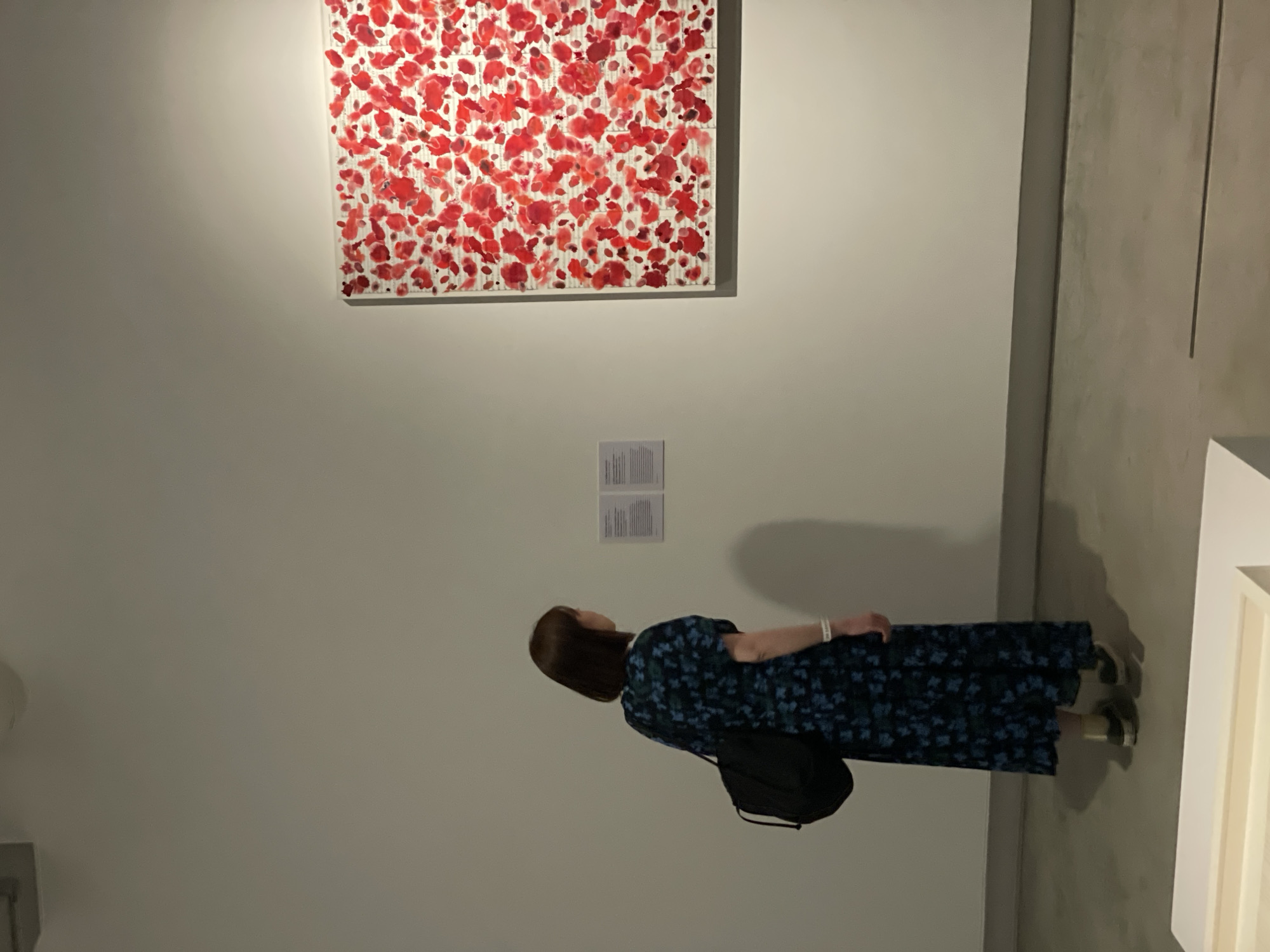 A woman stands in an art gallery, gazing up at a piece of artwork on the wall. The artwork, a square canvas, features an arrangement of red floral-like shapes against a white background. Below the artwork are two information cards. The woman, wearing a dark floral print dress and carrying a black bag, faces the artwork with her back to the viewer. The gallery is characterized by its clean, minimalist design, with white walls and neutral-colored floors. The right side of the image reveals a glimpse of another artwork on a white stand. The gallery is located in the city of Bangkok, Thailand.