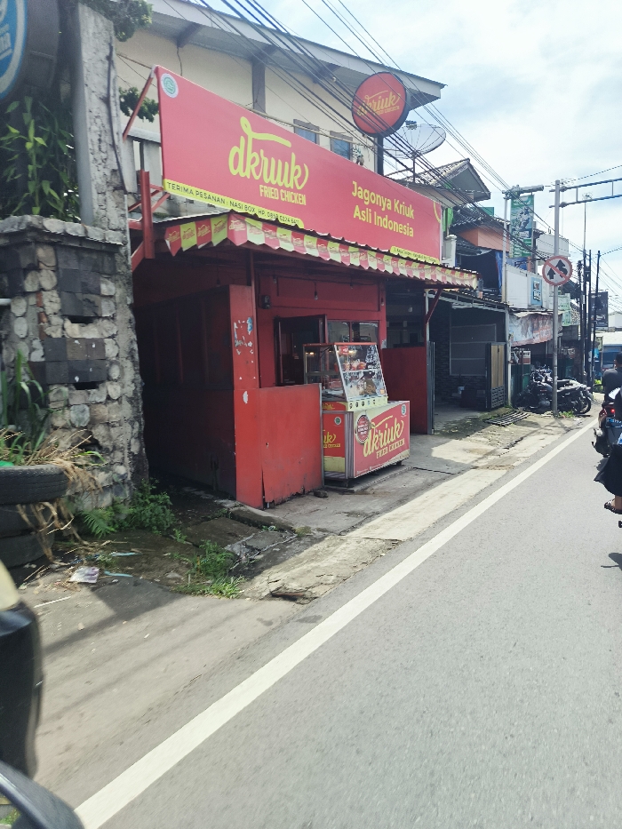 The image shows a street-side food stall, "D'kruk Fried Chicken," located at the coordinates -7.103, 107.461 in the city of Ciwidey, Indonesia. The stall is a small, red, brick building with a white awning and a red sign with the words "D'kruk Fried Chicken" and "Jagonya Krikuk Asli Indonesia". The stall has a window with a display of food and a glass-doored refrigerator. There are signs in Indonesian on the building. In front of the stall, a person is walking away from the camera, and a motorcycle is parked on the side of the road. There is a white line painted along the side of the road, which is asphalt and has a light gray color. There is a brick wall and plants next to the stall. The sky is blue with some white clouds. The image appears to be taken during the day, as there are shadows cast from the buildings.