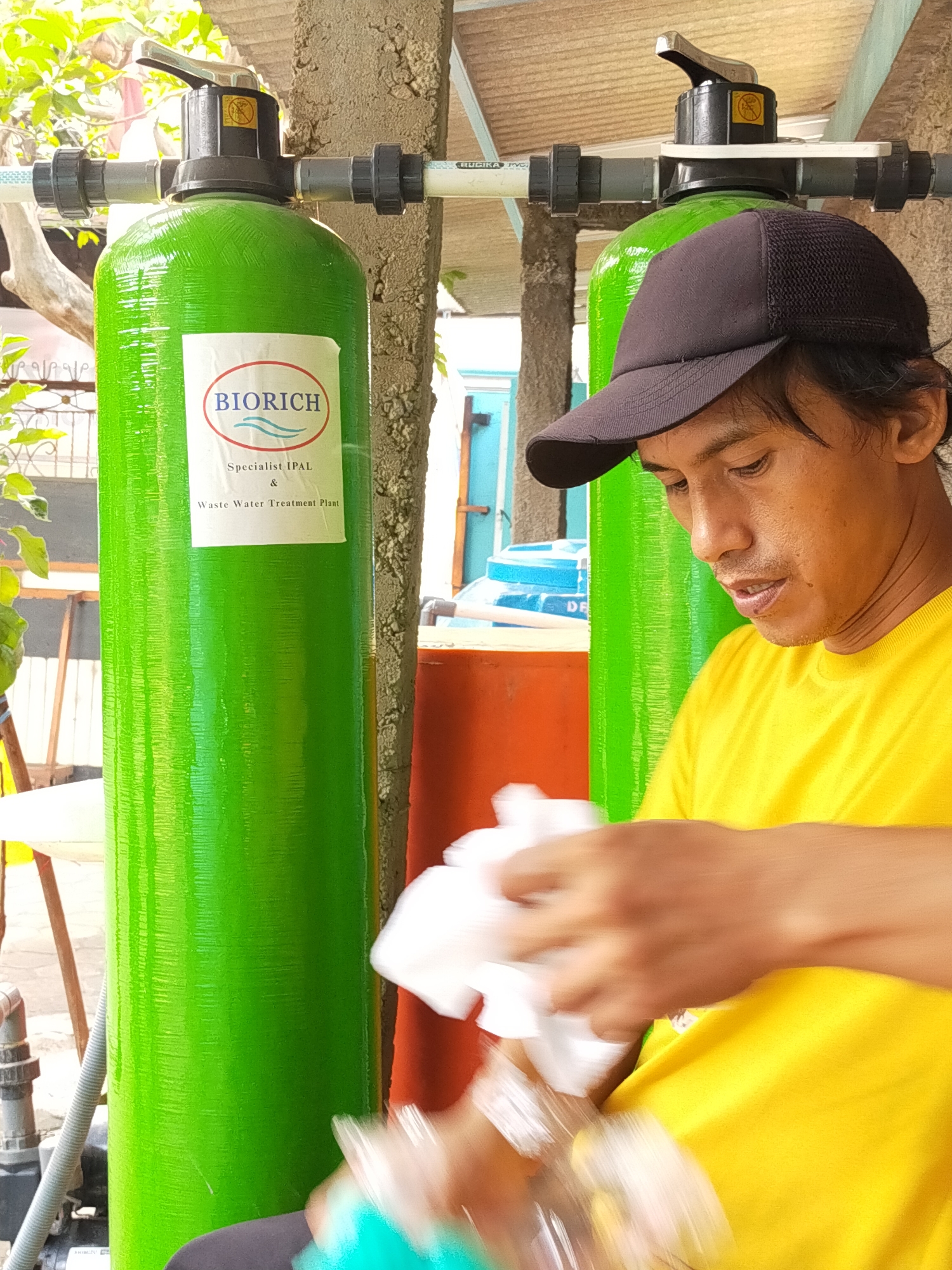 A medium shot captured outdoors in Tambun, Indonesia, depicts a man wearing a yellow t-shirt and a dark baseball cap, his head tilted downwards as he holds a white cloth and a blurry object in his hands. He stands to the right of two tall, bright green cylindrical tanks, each capped with black valves and connected by white PVC piping. A white label on the left tank reads "BIORICH Specialist IPAL & Waste Water Treatment Plant". The background reveals a textured concrete pillar, a partially visible orange barrel, and some foliage. The lighting suggests it is daytime, with sunlight illuminating the scene.