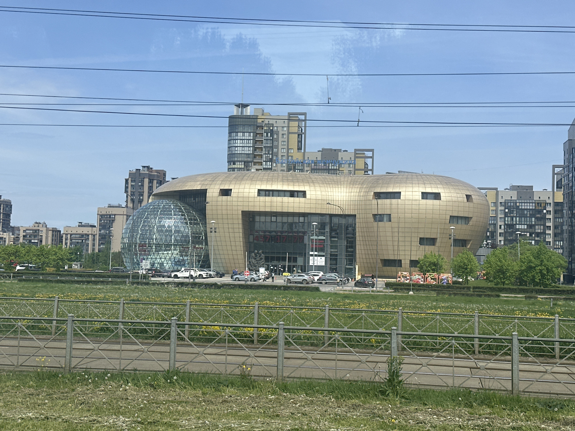 The image depicts a modern architectural complex in Saint Petersburg, Russia. The focal point is a large, bulbous building covered in golden-colored panels with a glass sphere attached to one side. The building has a central glass-walled entrance.
In front of the building is a grassy field dotted with yellow flowers and dandelions. A metal fence runs along the front of the field, separating it from a paved area or road. Beyond the building, multiple apartment buildings are visible against a blue sky with some scattered clouds. Power lines are strung across the upper portion of the frame.
Several cars are parked in front of the golden building. A few pedestrians are visible in the same area.