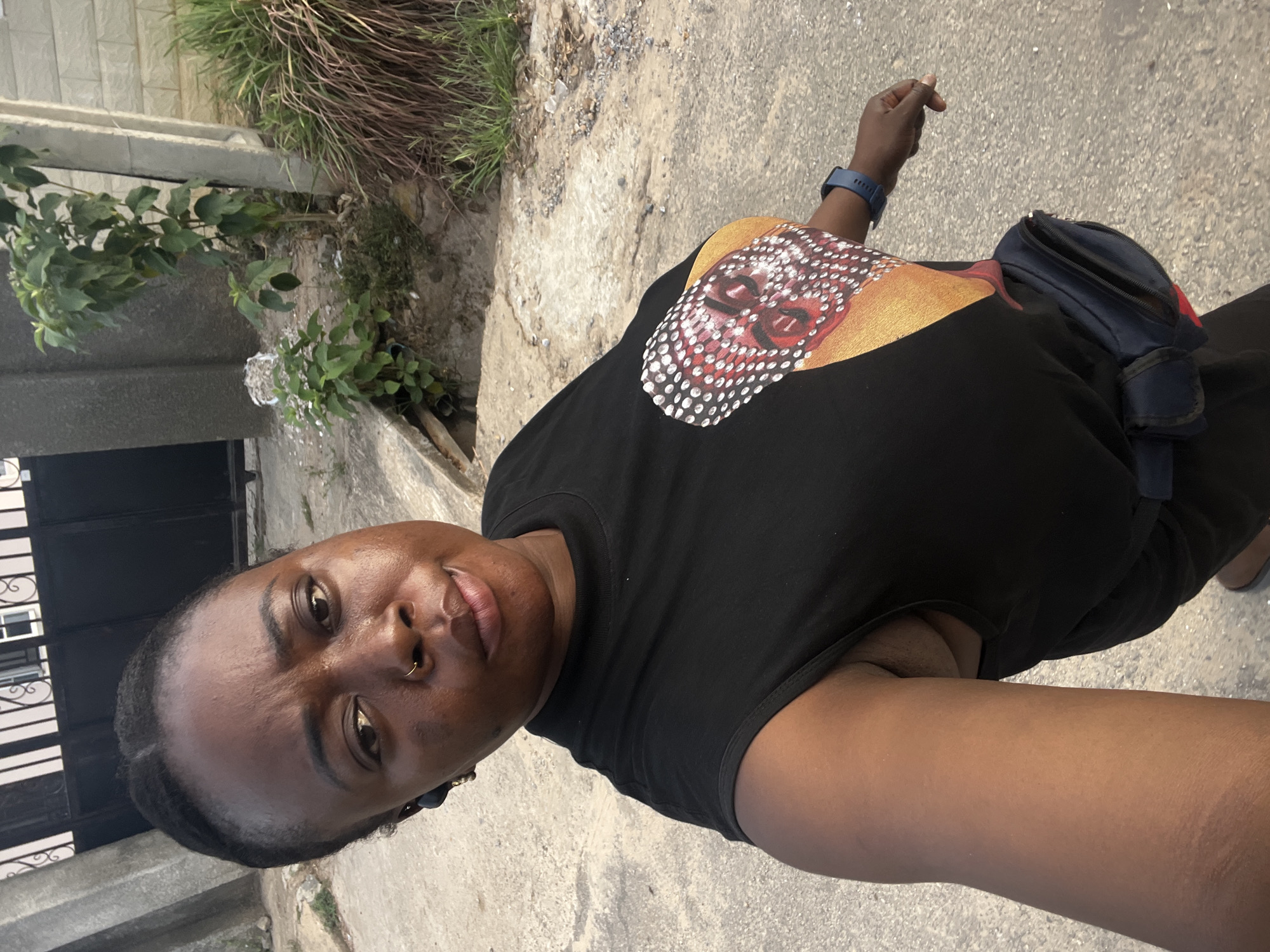 A person wearing a black t-shirt with a patterned design, a blue smartwatch, and a small dark blue bag slung across their body is standing on a concrete surface, possibly a sidewalk. The environment includes green plants and a gray wall to the left, and an asphalt or concrete surface with scattered debris underfoot. The picture seems to be taken in daylight. The person is facing the camera, likely in Abuja, Nigeria.