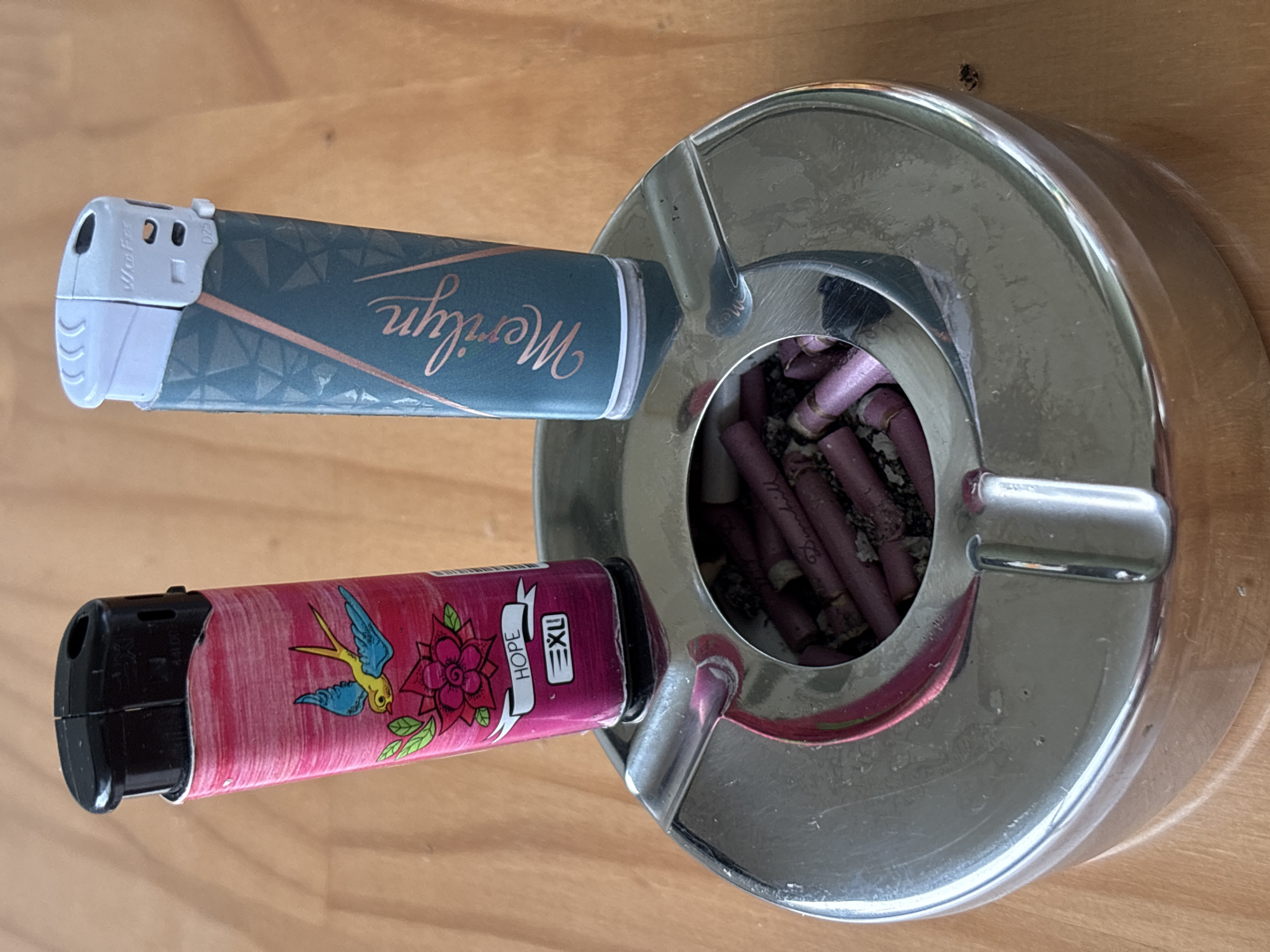 The image displays two disposable butane lighters resting within the indentations of a reflective metallic ashtray, which is situated on a light-colored wooden surface.
The lighter on the left features a pink, textured casing adorned with a light blue and yellow bird in flight above a stylized pink flower with green leaves. Below the flower, a white banner contains the word "HOPE" and the logo "EXU." Its top mechanism is black plastic.
The lighter on the right has a teal-colored casing with a subtle geometric pattern in a lighter, possibly rose-gold tone. The name "Marilyn" is written vertically in a script font on its side. Its top mechanism is light gray plastic, showing text partially legible as "Mer Fire" and "D25."
The ashtray is made of polished metal, likely chrome or stainless steel, exhibiting reflections of the lighters and ambient light. It has three U-shaped cigarette rests on its rim and a central circular opening. Inside this opening, numerous discarded cigarette butts with distinct dark pink or purple filters are visible, intermingled with ash and residual tobacco. The brand name "Dunhill" is clearly discernible on at least one of the cigarette filters.
The wooden surface beneath the ashtray shows a visible grain pattern, indicating a table or desk. The scene is illuminated by an unseen light source, creating highlights and reflections. No people are present in the image. No visual cues within the image indicate a specific city or country, including Plovdiv, Bulgaria.