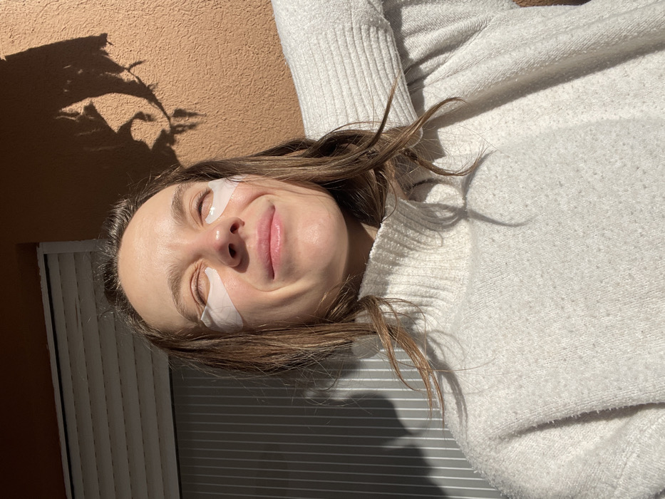A young adult female is depicted in a medium close-up, illuminated by direct sunlight, in an outdoor setting identified as Budva, Montenegro. The subject has her eyes closed and is smiling, with two white, triangular under-eye patches affixed beneath her eyes. She wears a white, knitted turtleneck sweater. Her brown hair is parted and falls across her shoulders. The background features a light orange textured wall on the right and a grey horizontal roller shutter on the left. Distinct shadows, including one resembling plant foliage, are cast upon the background elements. The subject's right arm is partially visible, bent at the elbow, extending out of frame to the right.