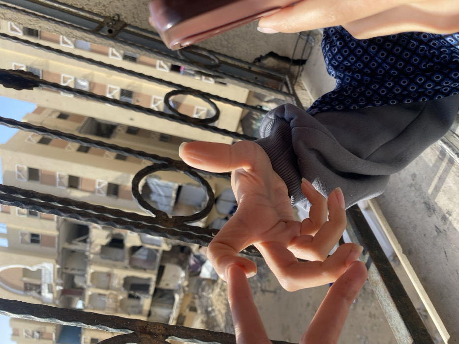 This image, captured in the city of Khan Yunis, Palestinian Territory, displays a scene viewed through dark, ornate metal railings. In the foreground, two hands are visible. The left hand, with manicured nails, performs an open gesture with its index finger and thumb extended. The right hand, clad in a grey sleeve cuff, executes a thumbs-up gesture while partially holding a maroon electronic device. Beyond the railings, a severely damaged multi-story building stands in the left midground, exhibiting significant structural collapse with large sections of its facade missing and internal structures exposed. To its right, a taller, multi-story residential building appears structurally intact. A clear blue sky forms the background.