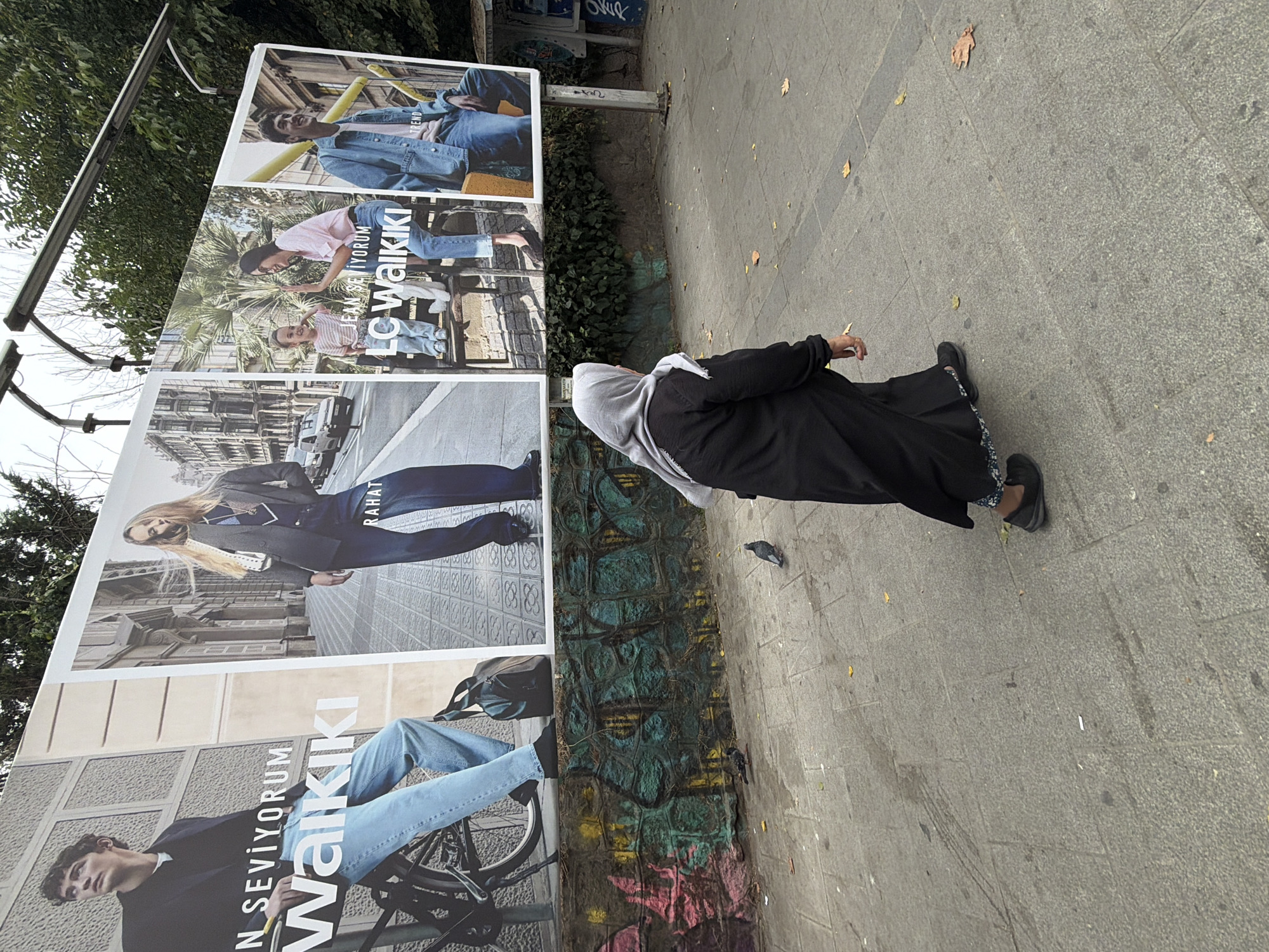 The image shows a street scene in Kadıköy, Turkey, with a large advertisement for LC Waikiki clothing brand displayed on a wall. In the foreground, a woman wearing a black abaya and a light gray hijab is walking away from the camera. She is on a paved sidewalk, and there are two pigeons on the ground near her. Behind the woman, the LC Waikiki advertisement features several models wearing casual clothing, with text in Turkish and English. The wall behind the advertisement is decorated with colorful graffiti. Trees and the sky are visible in the background. The overall environment suggests an urban outdoor setting.