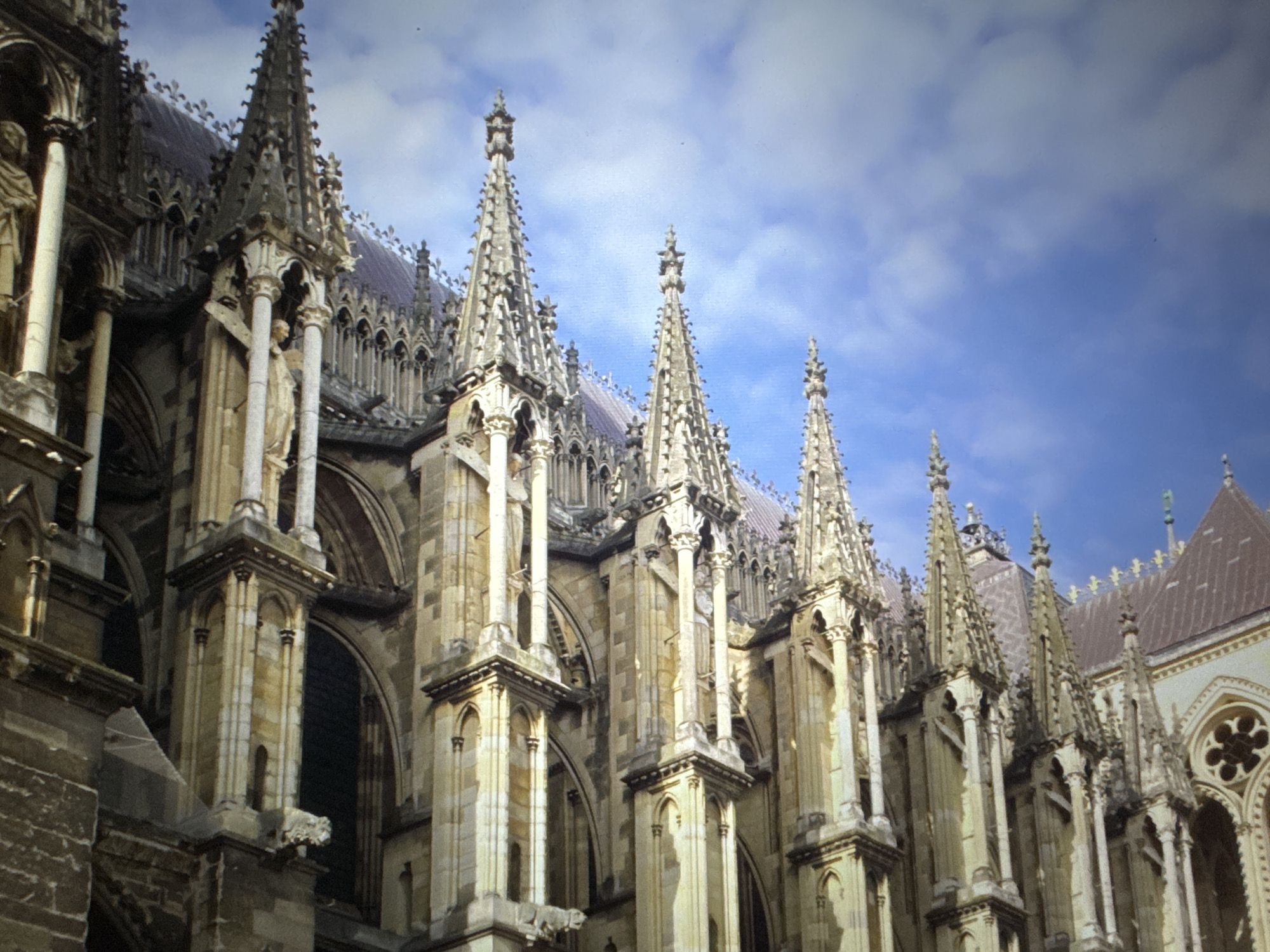 This media file presents a close-up view of the ornate exterior of a grand Gothic cathedral. The image is taken from a low angle, looking up towards the building's upper sections and roofline against a bright blue sky with scattered white clouds.
The primary subject is the cathedral's intricate architecture, characterized by numerous tall, pointed pinnacles (finials) that line the roof and adorn various architectural supports. These pinnacles are richly decorated with crockets and other carved details. Several flying buttresses are clearly visible, supporting the upper walls and adding to the structural and aesthetic complexity. The facade features large arched openings and windows, with detailed stone tracery. Sculptural elements are also present, including at least one discernible statue of a human figure within a niche and a grotesque gargoyle projecting from a lower section of the building. The stone used for construction appears to be a light, possibly beige or gray, material, which is well-lit by the daytime sun, creating distinct shadows and highlighting the texture of the carvings.
No specific activity is depicted, as the image focuses on the static architectural grandeur. The scene suggests a clear, sunny day. No people are visible, and there is no discernible text within the image. The overall impression is one of awe-inspiring craftsmanship and historical significance.