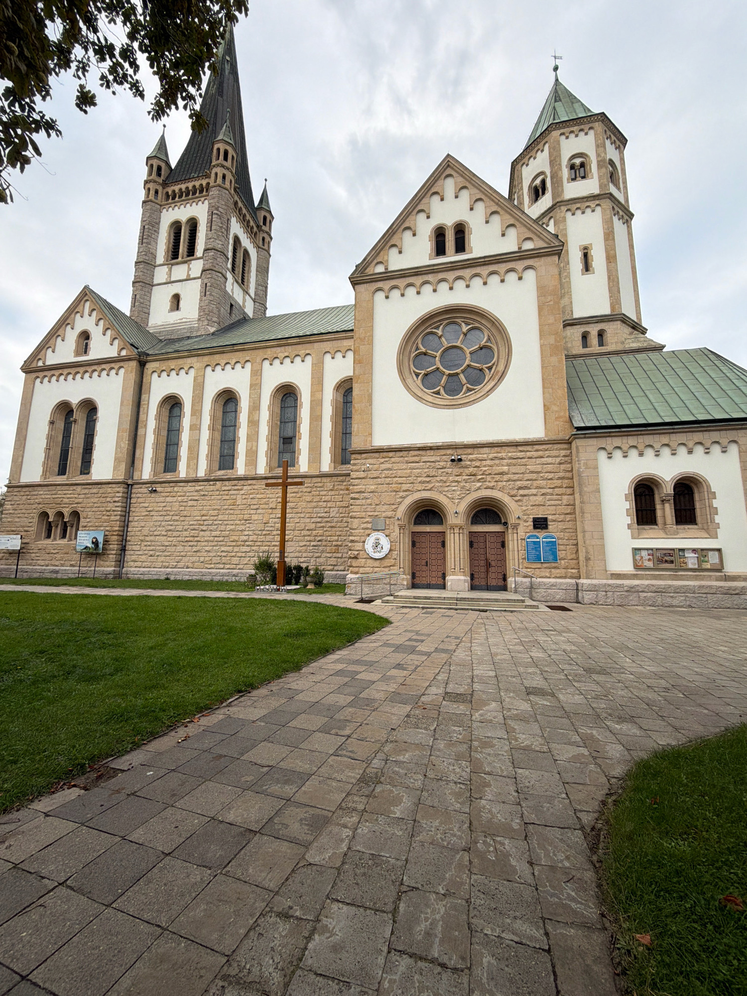 The image shows the Church of the Holy Name of Jesus in Wrocław, Poland. The church is made of stone and has a tall spire. In front of the church is a paved walkway and a grassy area. There is a wooden cross in front of the church. The sky is cloudy.
