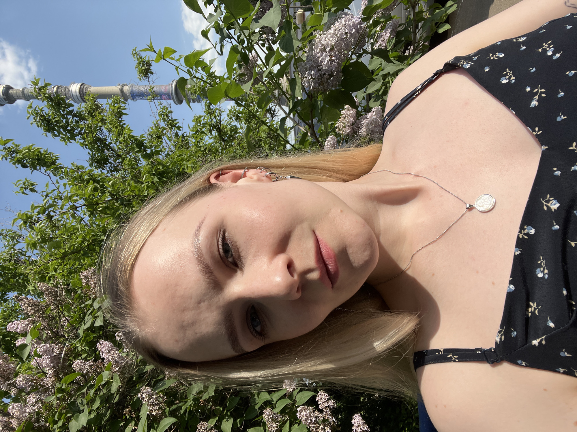 A fair-skinned woman with blonde hair is outdoors in Khimki, Russia, with the Ostankino Tower visible in the background. She is wearing a black floral dress and a silver necklace with a circular pendant. Lilac bushes surround her.