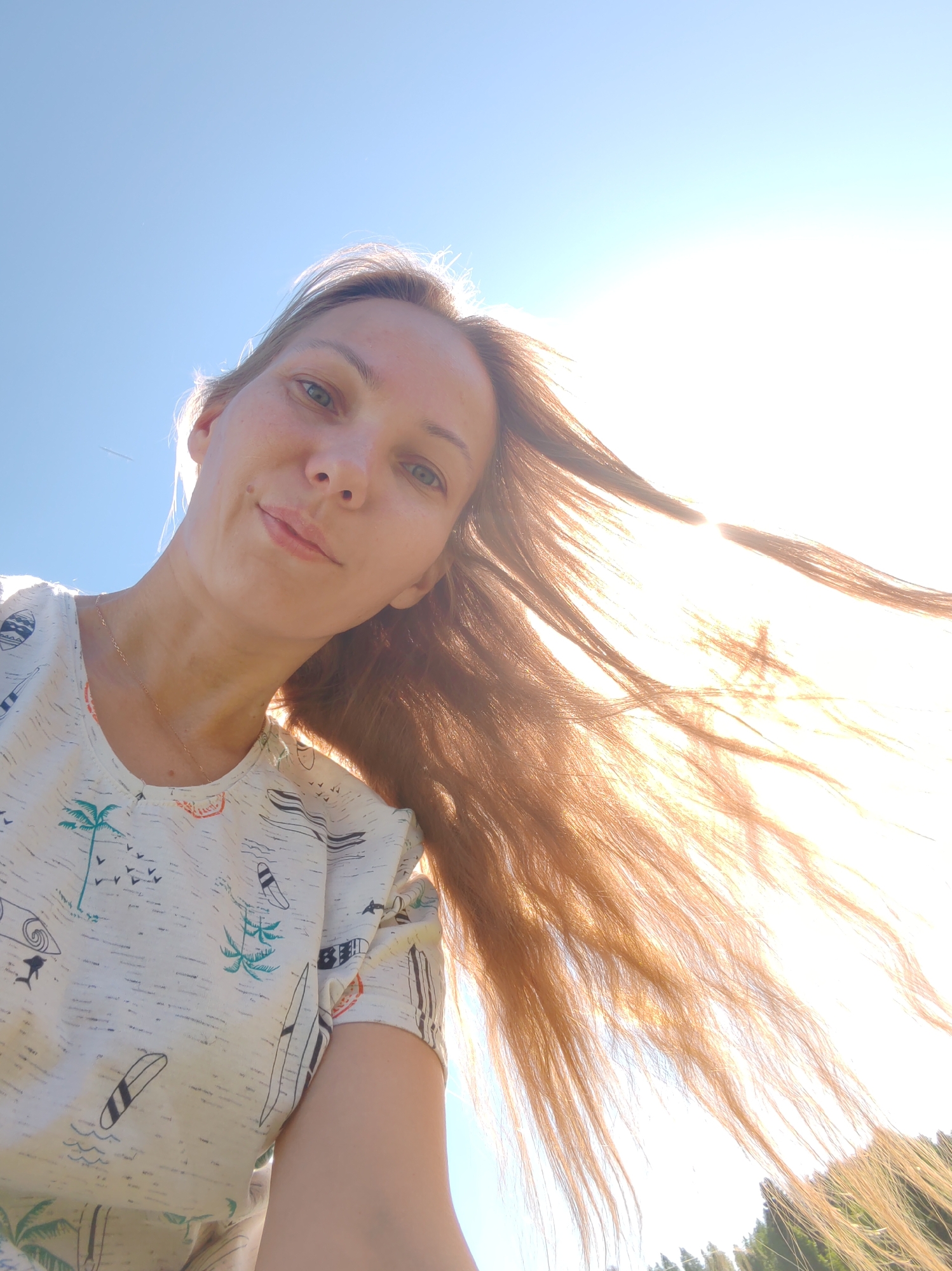 A low-angle selfie captures a fair-skinned woman outdoors under a bright, clear sky. She appears to be in her 20s or 30s, looking directly at the camera with a subtle smile. Her light-colored eyes are visible, along with scattered freckles on her face. Her long, light brown hair, illuminated by intense backlighting from the sun on the right, appears golden and is swept to the right, suggesting wind or motion. She wears a light-colored t-shirt patterned with beach-themed motifs, including palm trees, surfboards, and birds. A thin, golden necklace is visible on her neck. The upper left portion of the frame shows a blue sky with a faint aircraft contrail. The sun, positioned towards the upper right, creates significant overexposure and lens flare, making that area of the sky appear brilliant white. In the bottom right corner, the dark outlines of trees or foliage are faintly visible against the bright light. The scene is consistent with an outdoor setting on a sunny day in what is indicated to be the city of Чухлэм, Russia.