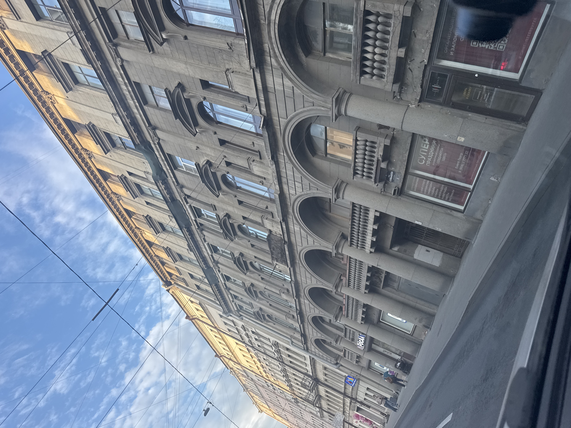 An angled street-level view captures a grand, multi-story building facade in Saint Petersburg, Russia, under a partially cloudy blue sky. The camera is tilted, looking upwards, with the building extending from the left foreground towards the right background. Overhead, various utility wires crisscross against the sky.
The building exhibits a classical or neo-classical architectural style, characterized by its grey stone appearance, numerous arched windows, decorative columns, and ornate balustrades. Sunlight illuminates the upper sections of the building, giving it a golden hue, while the lower parts are in shadow.
At street level, several commercial establishments are visible. Signs display Russian text, indicating services such as an "Event-Театр" (Event-Theater) that organizes "СУПЕР ПРАЗДНИКИ" (SUPER HOLIDAYS) including "КОРПОРАТИВЫ" (CORPORATE EVENTS), "СВАДЬБЫ" (WEDDINGS), "ЮБИЛЕЙ" (ANNIVERSARIES), "ДНИ РОЖДЕНИ" (BIRTHDAYS), "РОЗЫГРЫШИ" (RAFFLES), "КВЕСТЫ" (QUESTS), and "ФЕСТИВАЛИ" (FESTIVALS). Another sign promises "ОРГАНИЗАЦИЯ И ЗАПОМИН" (ORGANIZATION AND REMEMBER) along with "ИММЕРСИВНЫЕ ПРАЗДНИКИ" (IMMERSIVE HOLIDAYS) and a "ДВЕРЬ В СКАЗКУ" (DOOR TO A FAIRY TALE). A street sign with "КУЙБЫШЕВА" (KUIBYSHEVA) is also visible, hinting at the specific location within Saint Petersburg. The word "РОСАА" is also present on a different sign. A few indistinct figures can be seen walking on the sidewalk in the distance, suggesting daytime activity.