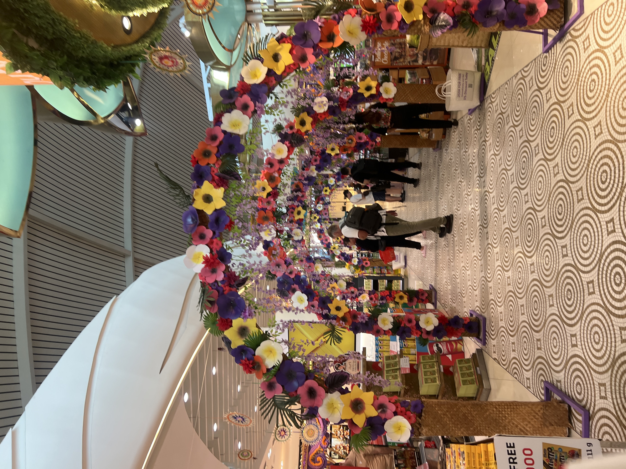 In Kuta, Indonesia, within a brightly lit indoor retail space, a decorative floral archway, composed of numerous artificial flowers in vibrant shades of purple, yellow, pink, and red, spans across a patterned tiled floor. The archway is supported by two tall, woven basket-like structures. Beyond the archway, people are visible navigating the aisles of a store, with shelves displaying various products, including chocolates and snacks. The ceiling features a modern design with white curved panels and black linear elements. In the foreground, a sign indicates a promotion with the word "FREE" and a monetary value. Further into the background, additional floral decorations and merchandise displays are present.