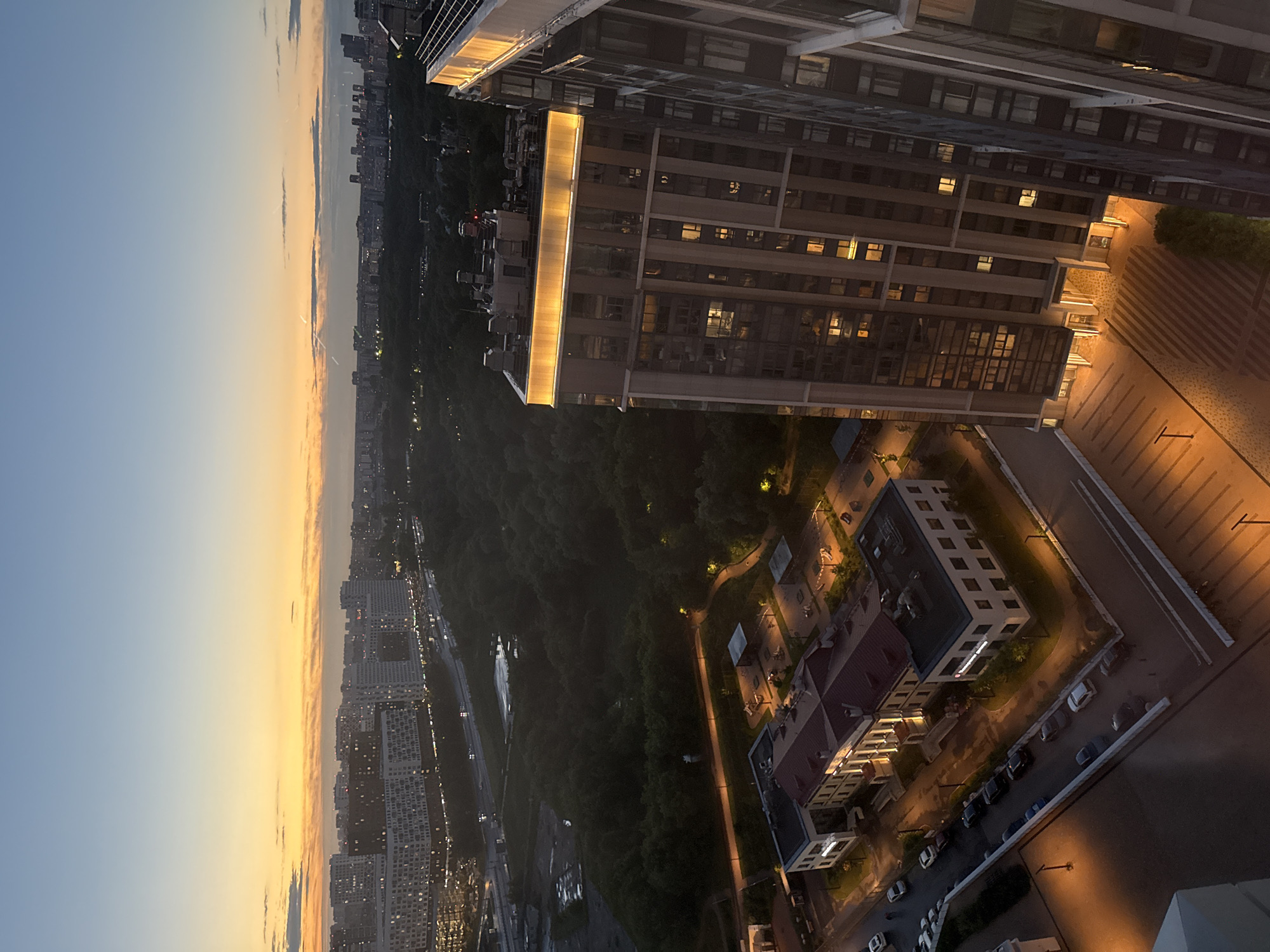 The image captures a twilight scene in Dolgoprudny, Russia, from a high vantage point, likely an apartment building. The sky displays a gradient of pale blue to warm orange near the horizon, indicating the sun has recently set or is about to rise.
In the foreground, a modern, tall residential building with many windows, some illuminated, dominates the right side of the frame. Its rooftop features decorative, warm-toned lighting. To the left and slightly behind this building, a complex of lower, rectangular buildings with red roofs is visible. These appear to be administrative or residential structures with illuminated windows. A street with parked cars, predominantly dark sedans and SUVs, runs alongside these buildings, and a few pedestrians are present on the sidewalks.
Further into the scene, a dense forest occupies a significant portion of the mid-ground, shrouded in shadow but with some pathways and streetlights visible, casting pools of yellow light. Beyond the forest, a sprawling cityscape extends to the horizon, characterized by numerous apartment buildings and infrastructure, including roads and what appears to be a highway with moving vehicle lights. The overall impression is an urban environment transitioning from day to night, with residential development coexisting with natural green spaces.