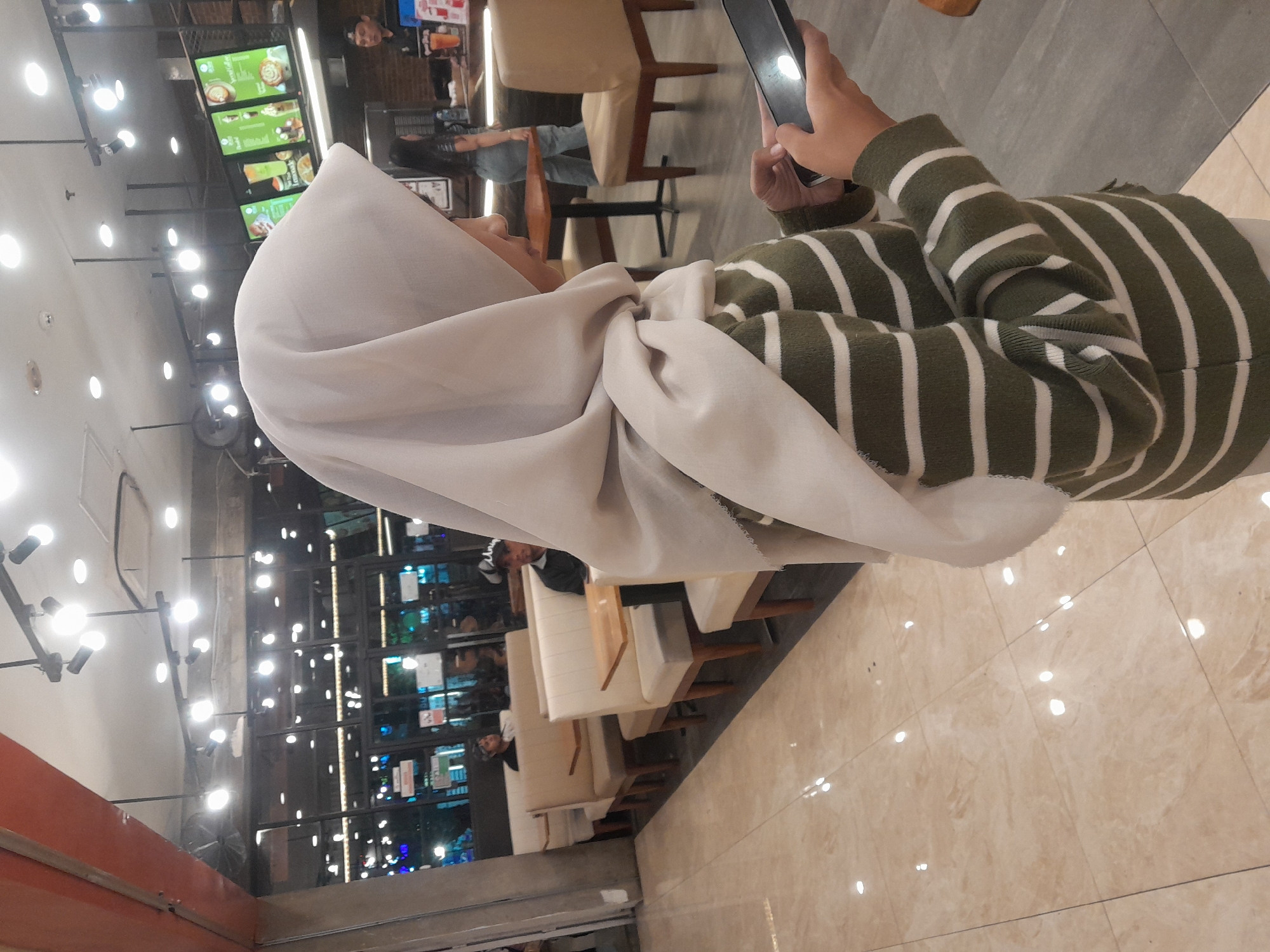 In a well-lit indoor restaurant, a young woman wearing a light-colored hijab and a green and white striped sweater is looking down at a smartphone in her hands. She is positioned in the foreground, slightly to the right of center, with her side profile visible. The restaurant's interior features beige upholstered booths with small wooden tables, tiled flooring that reflects the overhead lights, and a counter area in the background. Other patrons are seated in booths and at the counter. The ambiance suggests a casual dining establishment, possibly a fast-food or cafe setting. The image does not provide specific details that would allow for pinpointing its exact location within Banyuwangi, Indonesia, beyond it being an indoor dining establishment in that city.