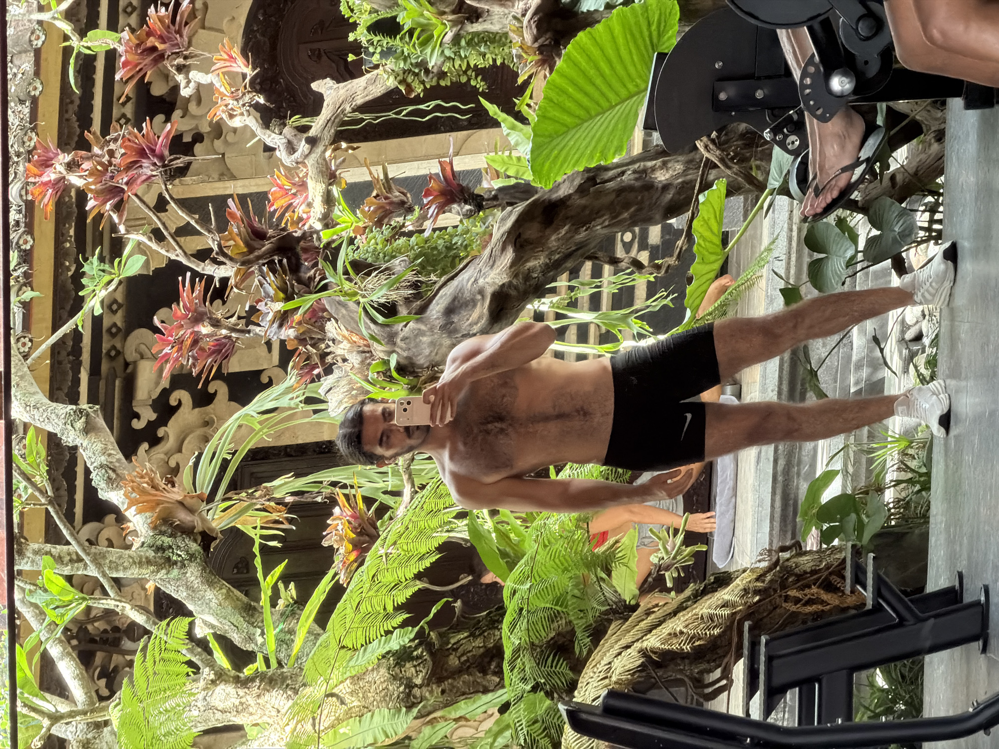 A shirtless man stands inside a gym near some plants and facing the camera. He is holding a phone in his left hand, which he is using to take a selfie. He is wearing black athletic shorts and white shoes. A small white logo is visible on the left side of his athletic shorts. Some gym equipment is visible on the upper right and lower portion of the image. On the left, is some intricate architecture with plants growing. The image was captured in North Kuta, Indonesia.