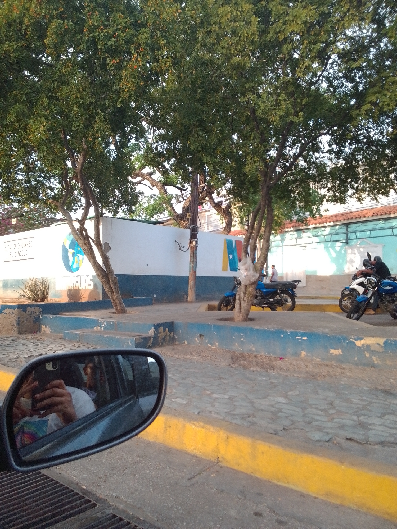 This image captures a street scene in Juan Griego, Venezuela, likely during the late afternoon given the warm, low sunlight casting long shadows. The setting appears to be an urban area with a building featuring a white facade and a blue lower section, possibly a public institution or business. Text on the building reads "ESTACION DE BOMBEROS EL CONC" and "AGUAS," suggesting a fire station and possibly a water-related facility.
In the foreground, a car's side mirror reflects a person taking a selfie. The person is wearing a white shirt and holding a smartphone. The mirror also shows the interior of the car, hinting at a journey in progress. Beyond the mirror, a gray metal grate is visible.
The scene is characterized by a large, leafy tree with a thick trunk, its branches reaching across the frame. The ground is paved with cobblestones and lined with a distinct yellow curb. Several motorcycles are parked or waiting on the side of the street. Two individuals are visible near the motorcycles; one person is seated on a white and blue motorcycle, and another, wearing a dark shirt, is adjusting something on a blue motorcycle. A single figure in a light shirt stands near the building.
The overall atmosphere is casual and everyday, with no indication of a specific event. The lighting suggests a pleasant, possibly warm, day. The presence of the fire station sign and the general urban environment provide clear location cues for Juan Griego.