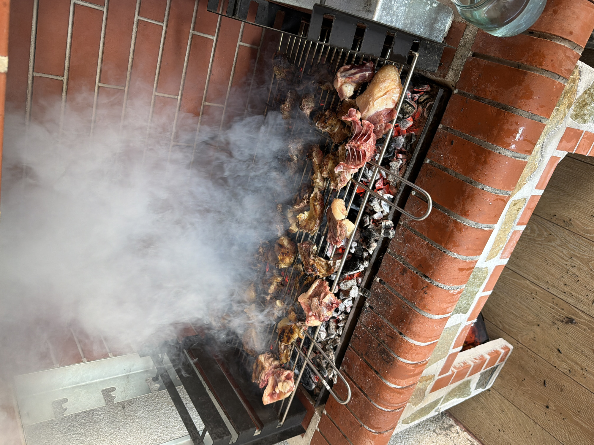 This image depicts a barbecue scene located in Sinitovo, Bulgaria. The primary subject is a large, brick-built outdoor grill, actively in use. Multiple pieces of raw and partially cooked meat, including cuts resembling chicken and various bone-in portions, are arranged on adjustable metal grates positioned over a bed of glowing charcoal and ash. A substantial volume of white smoke emanates from the cooking meat, rising and partially obscuring the upper left portion of the scene. The grill structure features reddish-brown bricks, with a curved top edge and a decorative pattern of mixed red, white, and pale green bricks on its lower left section. A red brick wall with visible mortar lines forms the background on the right. The ground immediately in front of the grill consists of light brown wooden floorboards. In the lower right foreground, a clear glass jar, partially filled with water, rests on the brick structure. No individuals are present in the image.