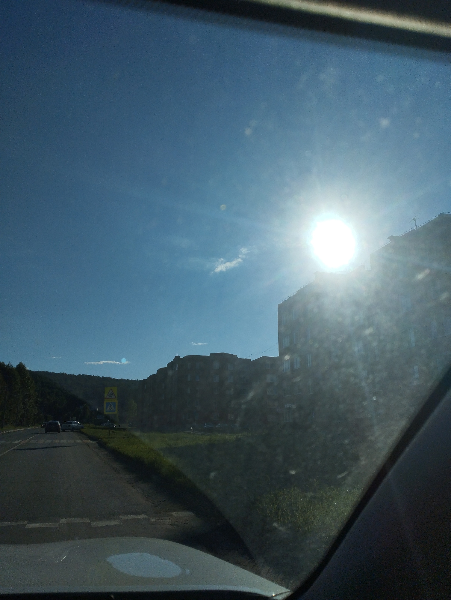 The image captures a view from inside a vehicle, looking out through the windshield. The scene is a residential area with apartment buildings situated along a road that curves to the left. A bright sun is visible in the sky, casting a glare and lens flare onto the image, suggesting it is daytime. The sky is blue with a few wispy clouds.
The road is paved and has white dashed lines indicating lanes. There are at least two cars on the road, moving away from the viewer. On the right side of the road, there is a grassy area and a row of apartment buildings. The buildings appear to be multi-story residential structures. On the left side of the road, there is a densely wooded hillside.
Two road signs are visible near the left side of the road. One sign is yellow and triangular with a black symbol, likely indicating a hazard or caution. Below it is a rectangular sign with a black pedestrian symbol, indicating a crosswalk. The presence of these signs and the apartment buildings suggest an urban or suburban environment. The overall mood is tranquil, with the bright sunlight creating a sense of a pleasant day. The time of day is clearly daytime, given the intense sun. The weather appears to be clear and sunny.