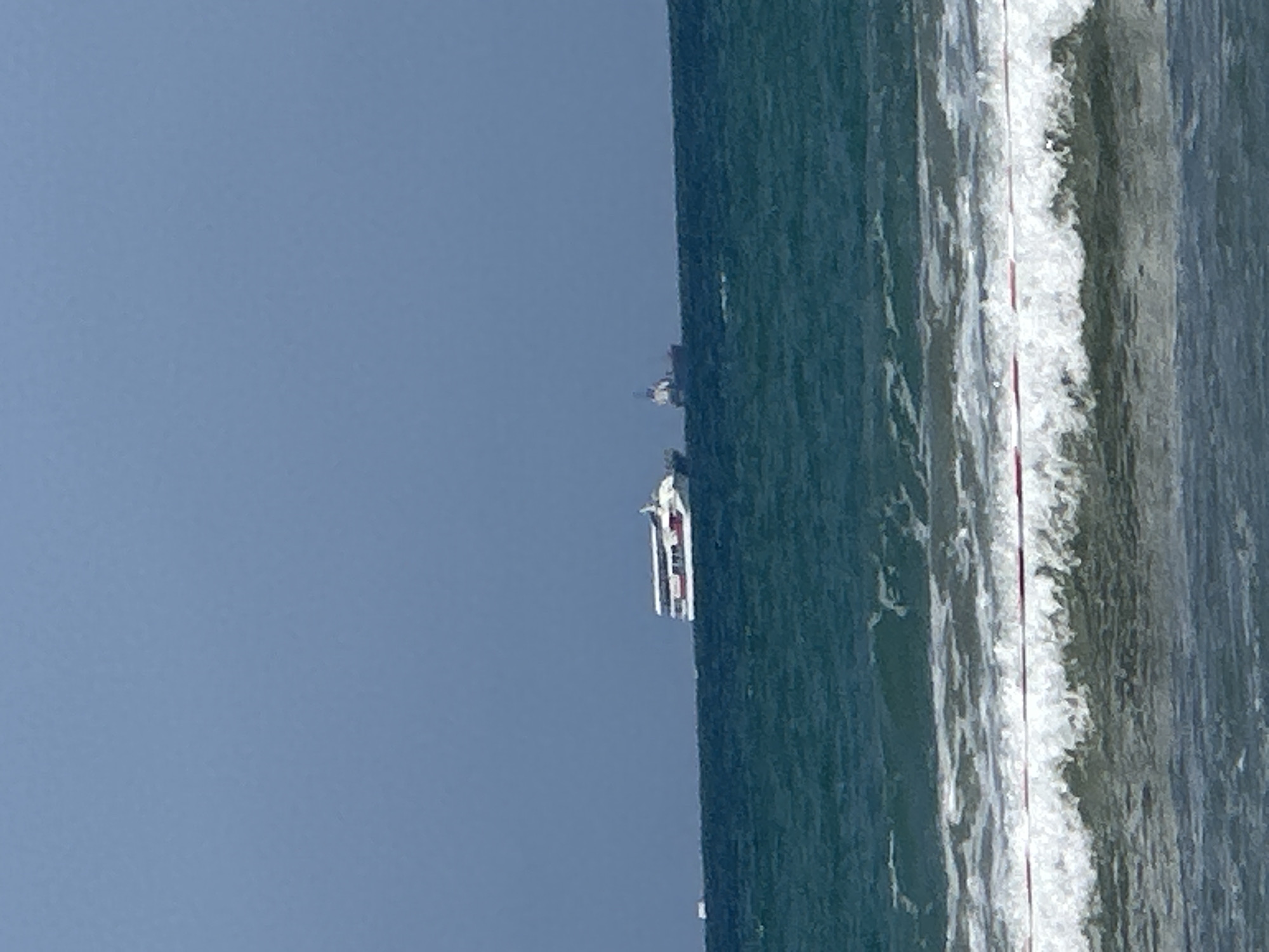 The image shows a view of the Mediterranean Sea from the shore in Kiryat Yam, Israel. In the distance, two vessels are visible on the water. The closer vessel is a white and red catamaran or ferry, with multiple decks and windows. Further to the right and slightly behind it, a smaller, darker boat with a visible superstructure is present. The sea is choppy with visible waves breaking near the shore. A thin, horizontal, red and white line, possibly part of a buoy or marker, is visible across the foreground waves. The sky is a clear, pale blue.