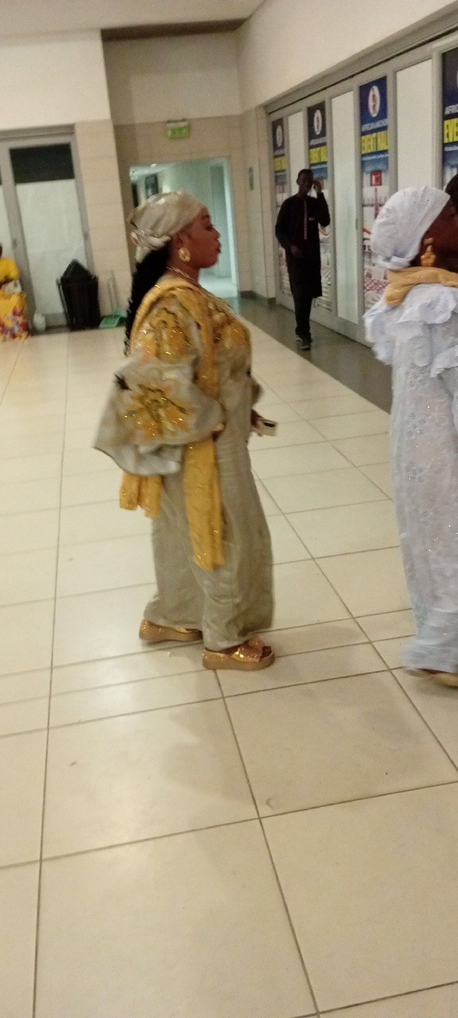 An undated, medium shot, side-profile image shows a woman with dark hair and dark skin wearing a light gray headscarf, large gold hoop earrings, and a light gray, floor-length outfit adorned with gold embroidery and embellishments. A yellow scarf is draped around her shoulders and neck, and she wears gold platform sandals. The woman is standing on a light-colored tiled floor, and behind her, a man in a dark suit walks away from the camera. In the background, doors are visible with posters indicating "EVENT HALL." The setting appears to be an indoor event space or convention center. The image does not provide specific location details for Gandu Albasa, Nigeria.