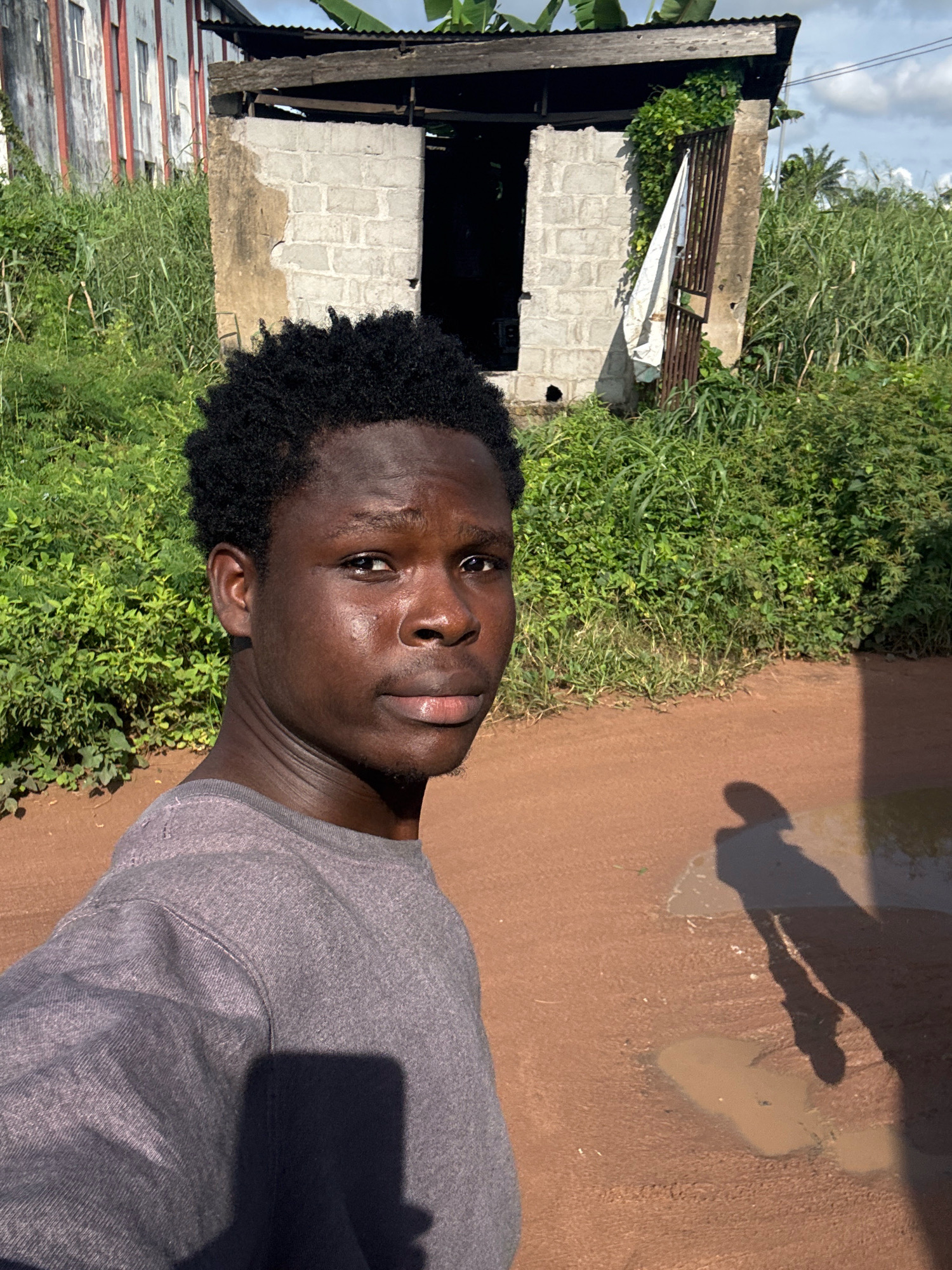 In the city of Ihiagwa, Nigeria, a young Black male is pictured outdoors, standing in the foreground and facing the camera. He has dark, curly hair and is wearing a gray crew-neck t-shirt. The background features a dilapidated structure with a corrugated metal roof and concrete block walls, partially covered in vines. To the left of this structure is a tall, white building with red trim and visible windows. Dense green vegetation surrounds the scene, and the ground is a reddish-brown dirt surface with puddles. A shadow of the male is cast on the ground to his right.