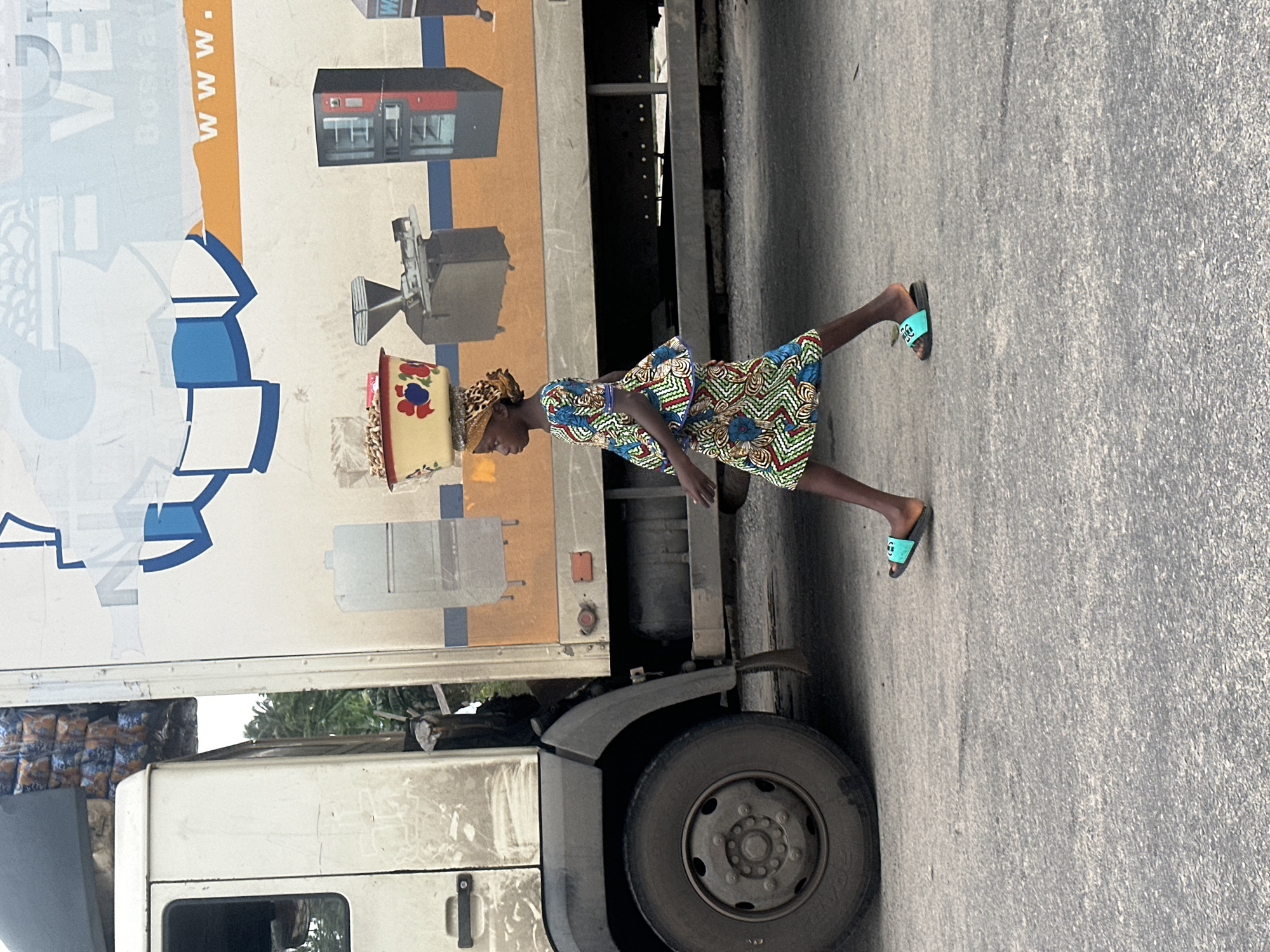 A young girl, likely a vendor, walks on a paved road beside a white truck in Badagry, Nigeria. She is wearing a vibrantly patterned dress and a headscarf. Balanced on her head is a large bowl filled with what appears to be nuts or seeds, and on her feet, she wears bright blue slide sandals. The truck has a white cab with a black door handle and a large, dirty tire. Its side displays a graphic mural featuring industrial equipment, including a refrigerator, a grinder, and possibly a vending machine, with text in English such as "Boskan" and "www.". The background shows foliage, indicating an outdoor urban setting.