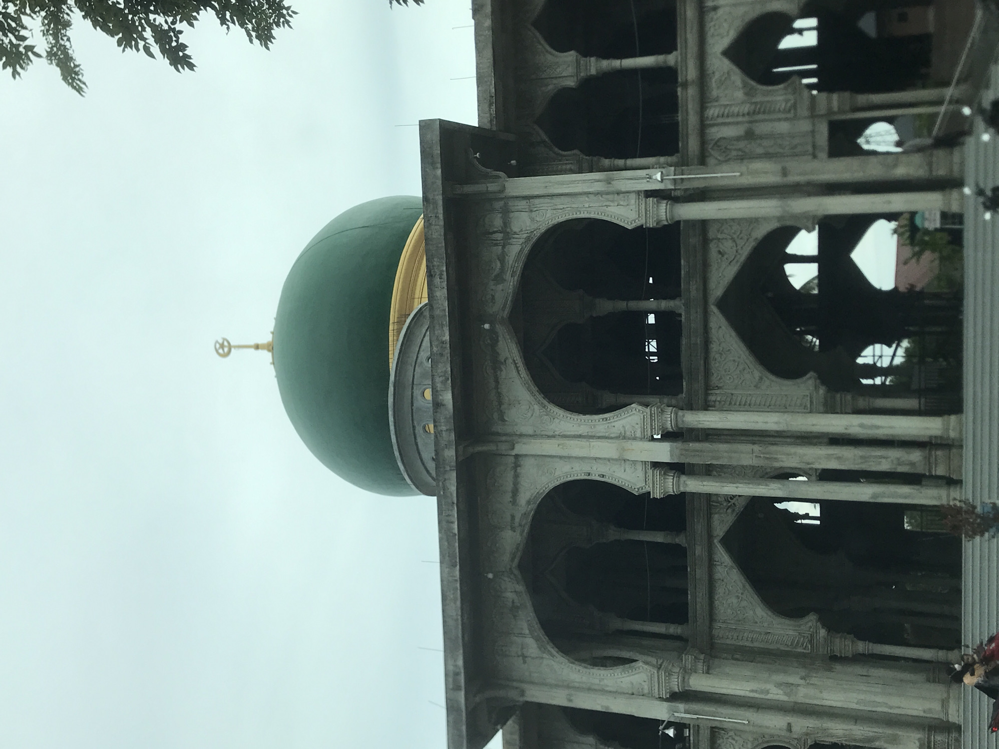 The image shows a mosque in Keudealue Ie Puteh, Indonesia. It features a green dome with a golden crescent and star on top. The building has multiple levels with arched openings supported by pillars and decorated with intricate carvings. The sky is overcast, and a tree branch is visible in the upper left corner of the frame. People are faintly visible in the lower right, outside the main structure.