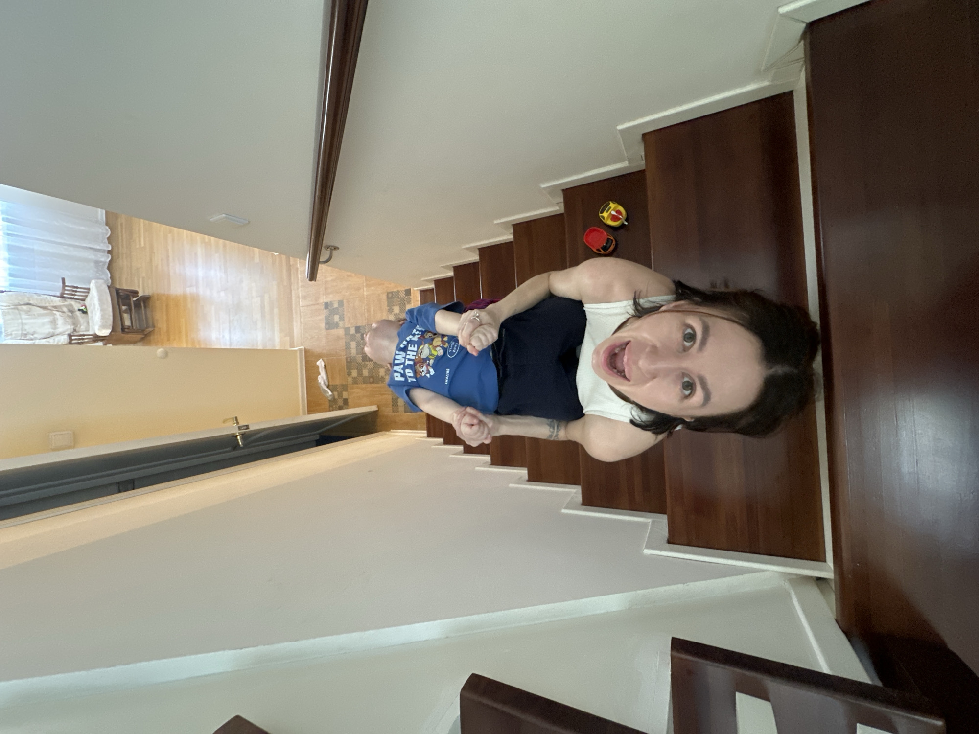 A woman is standing on a staircase holding a young child's hand, both facing the camera. The woman is wearing a white tank top and dark shorts, with dark hair. The child is wearing a blue Paw Patrol shirt. The staircase has dark wood treads and white risers, leading down into a light-colored room with wooden floors and furniture. The location is presumably in a residential building in Русский Вожой, Russia. There are toys visible on the stairs. The woman is looking at the camera and appears to be talking or yelling.