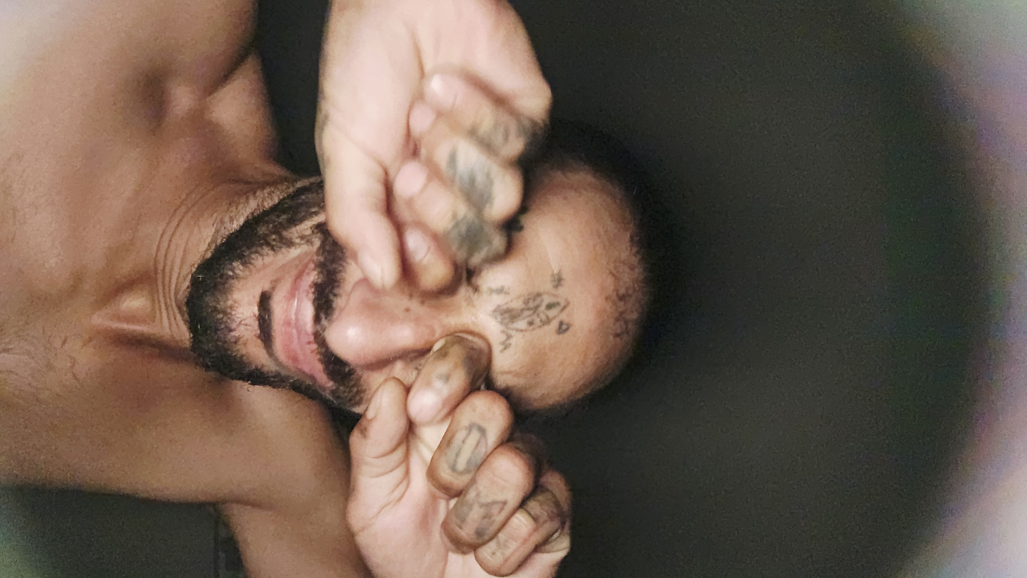 The image presents a close-up, rotated portrait of a person, likely male, from the chest up. The person's head is tilted and rotated, with their face partially obscured by both hands. Fingers are pressed against the forehead, covering the eyes, while the other hand covers part of the nose and mouth. The mouth is slightly open, revealing teeth. The person has dark facial hair, forming a beard, and short hair on their head. Faded tattoos are visible on the forehead, including a small, symbolic design and what appears to be the letter "W". Additional tattoos, comprising indistinct, abstract shapes or letters, are present on the fingers. The background is uniformly dark and out of focus, suggesting an indoor setting with low light. The lighting on the person's skin is somewhat harsh, highlighting facial features and hands. The gesture of covering the face conveys a sense of distress, weariness, or deep thought. The provided location context for this media file is Cam Đức, Vietnam.