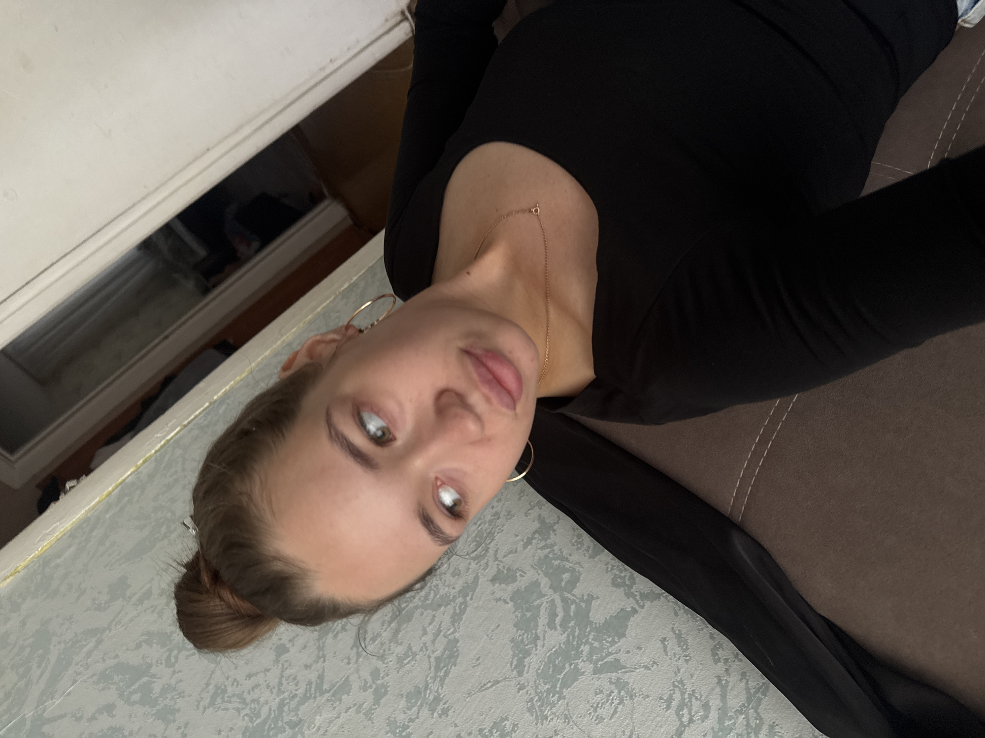 The image shows a young woman with brown hair styled in a bun, wearing a black long-sleeved shirt and a thin gold necklace. She is wearing large hoop earrings. Her head is turned to her left, and she is looking away from the camera with a slightly pursed expression. The background appears to be an indoor setting with a textured wallpaper in shades of blue and gray, and a brown couch or upholstered surface. A doorway or partition is visible to the right, showing a glimpse of a darker interior space with some objects. The lighting suggests it might be daytime. The specific location in Chaikovsky, Russia, cannot be determined from the visual information alone.