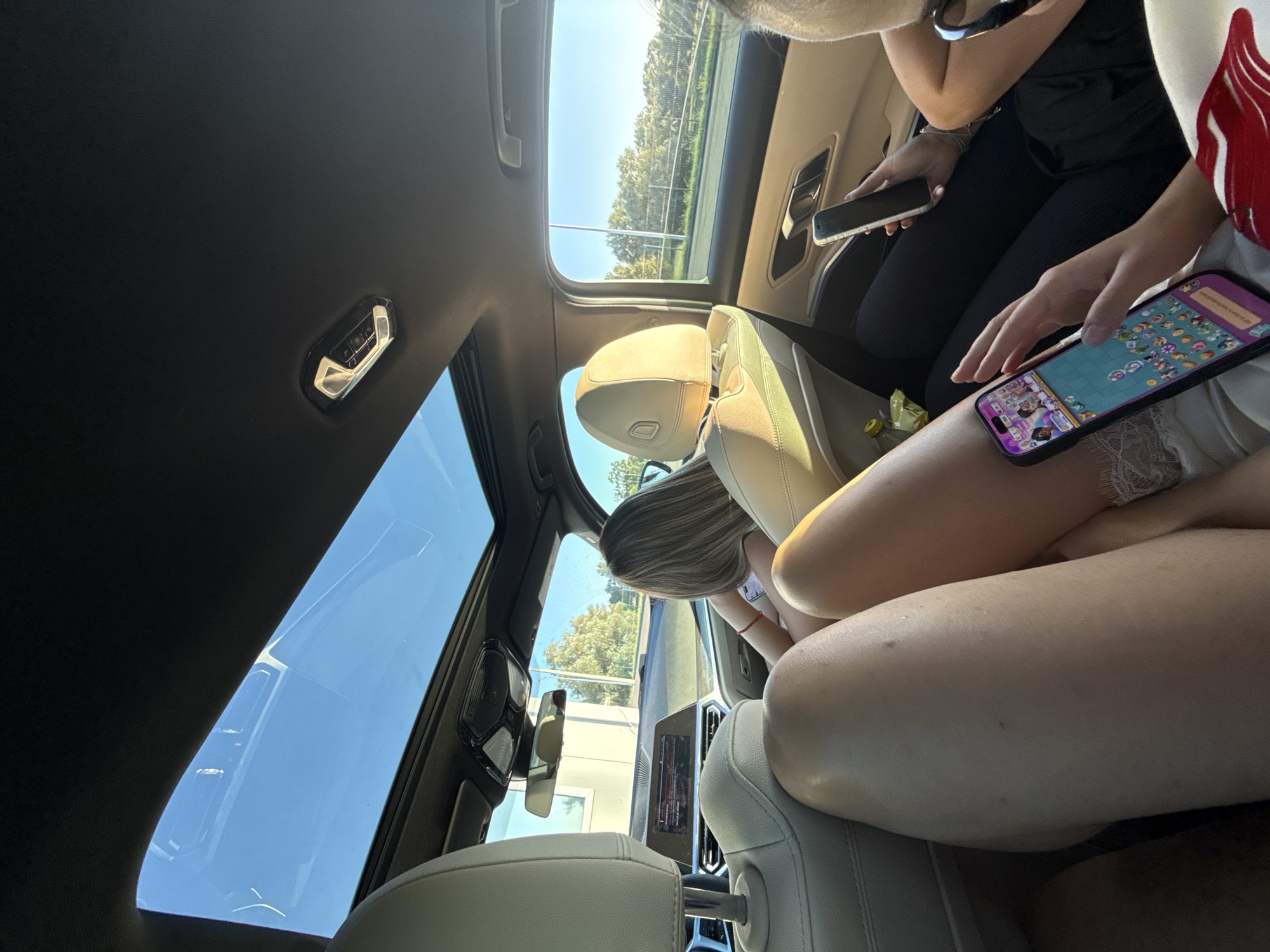 An interior shot captures the rear of a car's passenger cabin, focusing on three individuals. A person in the foreground has their legs positioned in the lower half of the frame, with one leg displaying a glimpse of white lace trim at the hem of their garment. To the right, another individual is seated, holding a smartphone and engaged in playing a game displayed on its screen. A third person is visible further back, also holding a smartphone, though their activity is not discernible. The car's interior features beige leather seats and a sunroof that reveals a bright blue sky. The exterior view through the windows shows trees and a road, suggesting a journey in a semi-urban or rural setting. The overall scene depicts a casual moment during a car ride.