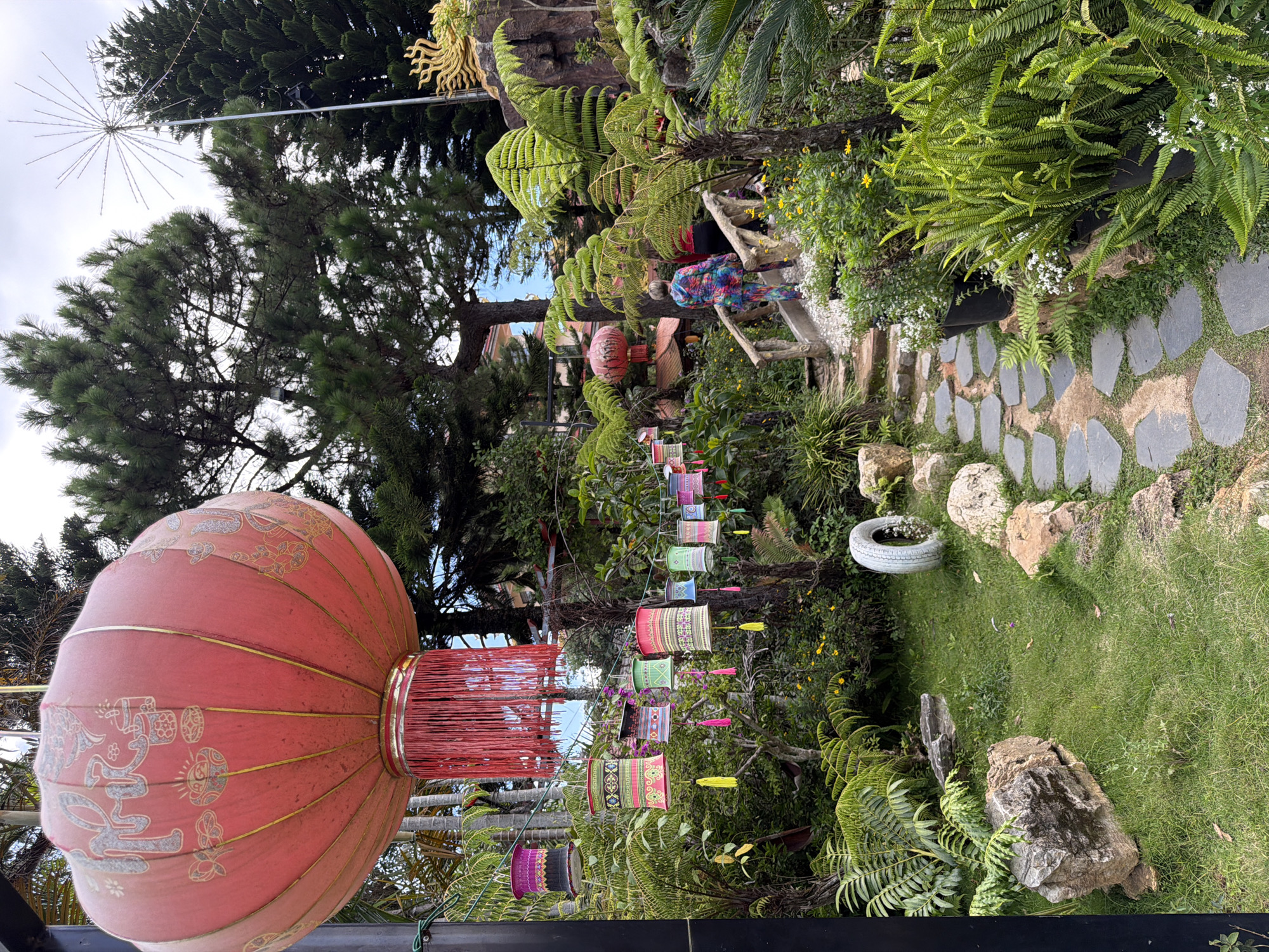 The image depicts a serene garden scene in Lâm Viên - Đà Lạt, Vietnam. In the foreground, a large red lantern with intricate golden Chinese characters hangs from a dark pole, adorned with a vibrant red fringe. To the left, a line of smaller, colorful lanterns, decorated with traditional patterns and tassels, are strung along a wire. A winding path of irregular grey stones leads through lush green grass and ferns, bordered by various plants and rocks. In the mid-ground, a wooden bridge spans a small incline, with two individuals walking across it. The person in the front is wearing a brightly colored, patterned outfit, and the person behind them is wearing red. Further back, red lanterns are visible amongst the trees, and a large, ornate golden sculpture of a sun or starburst is perched on a rocky outcrop. Tall pine trees dominate the background, with a cloudy sky overhead. The overall impression is one of a peaceful, well-maintained garden space, possibly a temple or tourist attraction, decorated with traditional cultural elements.