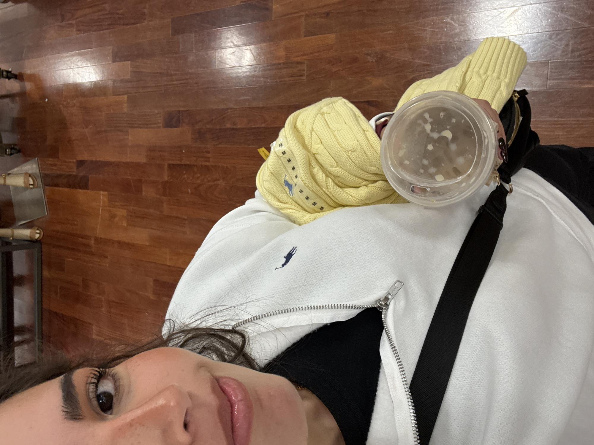 A close-up, slightly angled photograph depicts a person holding a clear plastic container with a translucent liquid and some white solids inside. They are wearing a white zip-up hooded sweatshirt with a Polo Ralph Lauren logo, and a black shirt underneath. A yellow knit sweater is draped on the person's shoulder, and a black bag strap is visible. The background shows a polished wooden floor. The city of San Marcos, United States of America is not able to be ascertained with the given image and information.