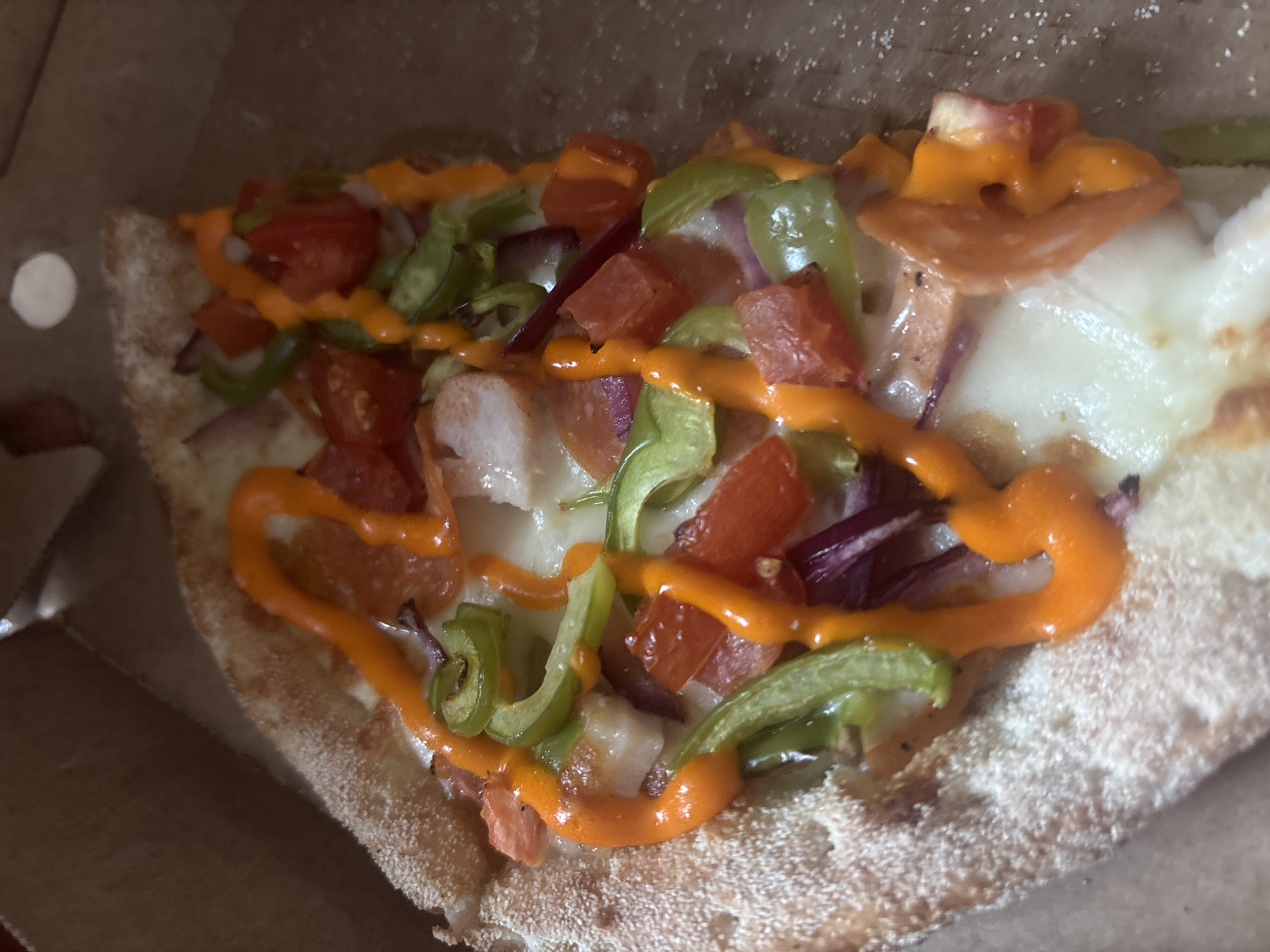 A slice of pizza is presented in a cardboard pizza box. The pizza has a thick, golden-brown crust that is dusted with flour. The toppings include melted white cheese, chunks of tomato, sliced green bell peppers, red onion, and what appears to be pieces of cooked chicken or sausage. A vibrant orange sauce is drizzled in a wavy pattern over the toppings. The background is the brown interior of the pizza box, with some crumbs visible. The scene is captured from a top-down perspective. There is no indication of the city of Aktobe, Kazakhstan, or any other specific location.