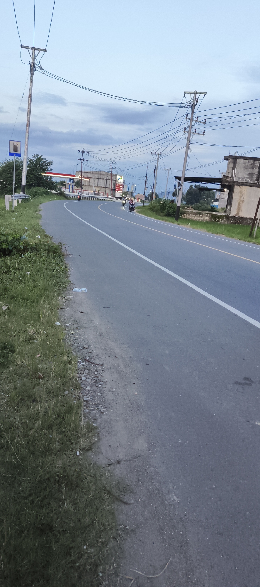 The image depicts a curving asphalt road in Bateiiliek, Indonesia, marked with a solid white line along its center. The roadside is covered with green grass and patches of small rocks and debris. A gas station with a prominent red sign is visible in the distance beyond a short concrete barrier. Several motorcycles are traveling on the road. Utility poles with attached power lines line both sides of the road. A fuel station sign is on a pole on the left side of the road. A partially constructed or dilapidated building is visible on the right side. The sky overhead is overcast.