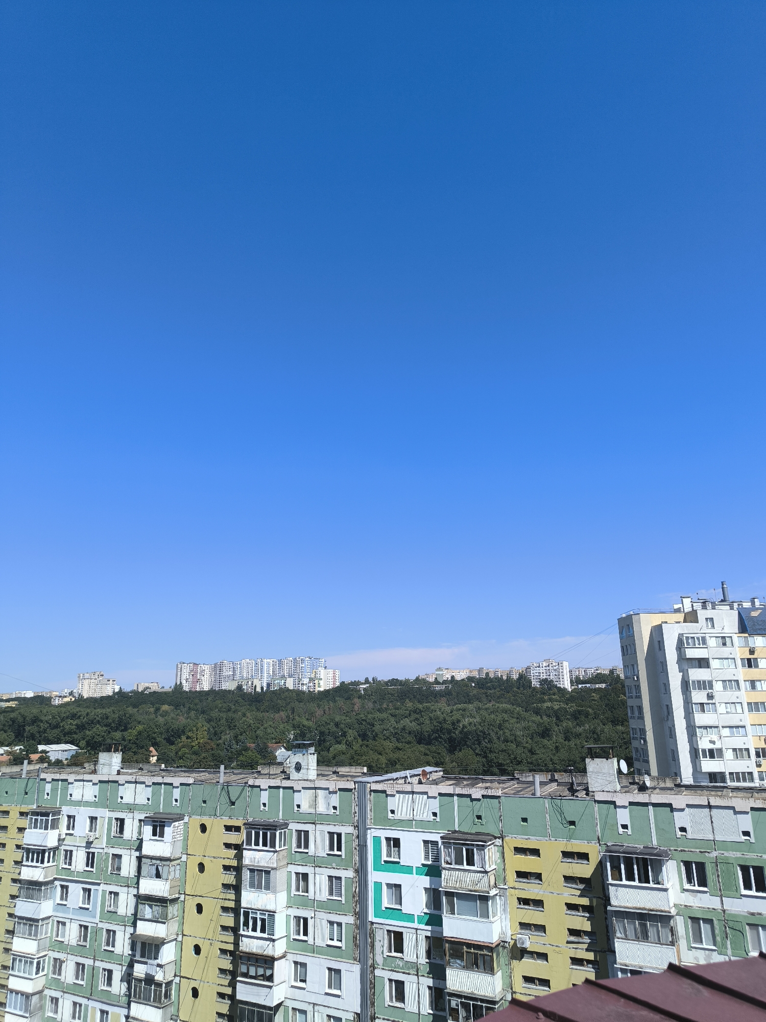 The image shows a view of residential buildings in Chișinău, Moldova, on a clear, sunny day. The foreground features a section of a multi-story apartment building with balconies. This building is painted in alternating sections of green and yellow with some light blue accents. It appears to be an older, Soviet-era style apartment block, characterized by its uniform design and practical architecture.
In the middle ground, a lush green, densely wooded hillside rises, dotted with more apartment buildings, some of which are much larger and more modern-looking than those in the foreground, with many windows. The trees suggest a natural landscape integrated with urban development.
In the background, further apartment buildings are visible on the crest of the hill, appearing as a cluster of white structures. To the right of the foreground building, a taller, modern apartment building is visible, painted in a light grey and yellow color scheme, with a more contemporary design featuring large windows and balconies.
The sky is a vibrant, cloudless blue, indicating fair weather. The overall scene depicts a segment of an urban environment in Chișinău, where older residential blocks are situated near a green hillside topped with newer constructions, under a bright, clear sky.