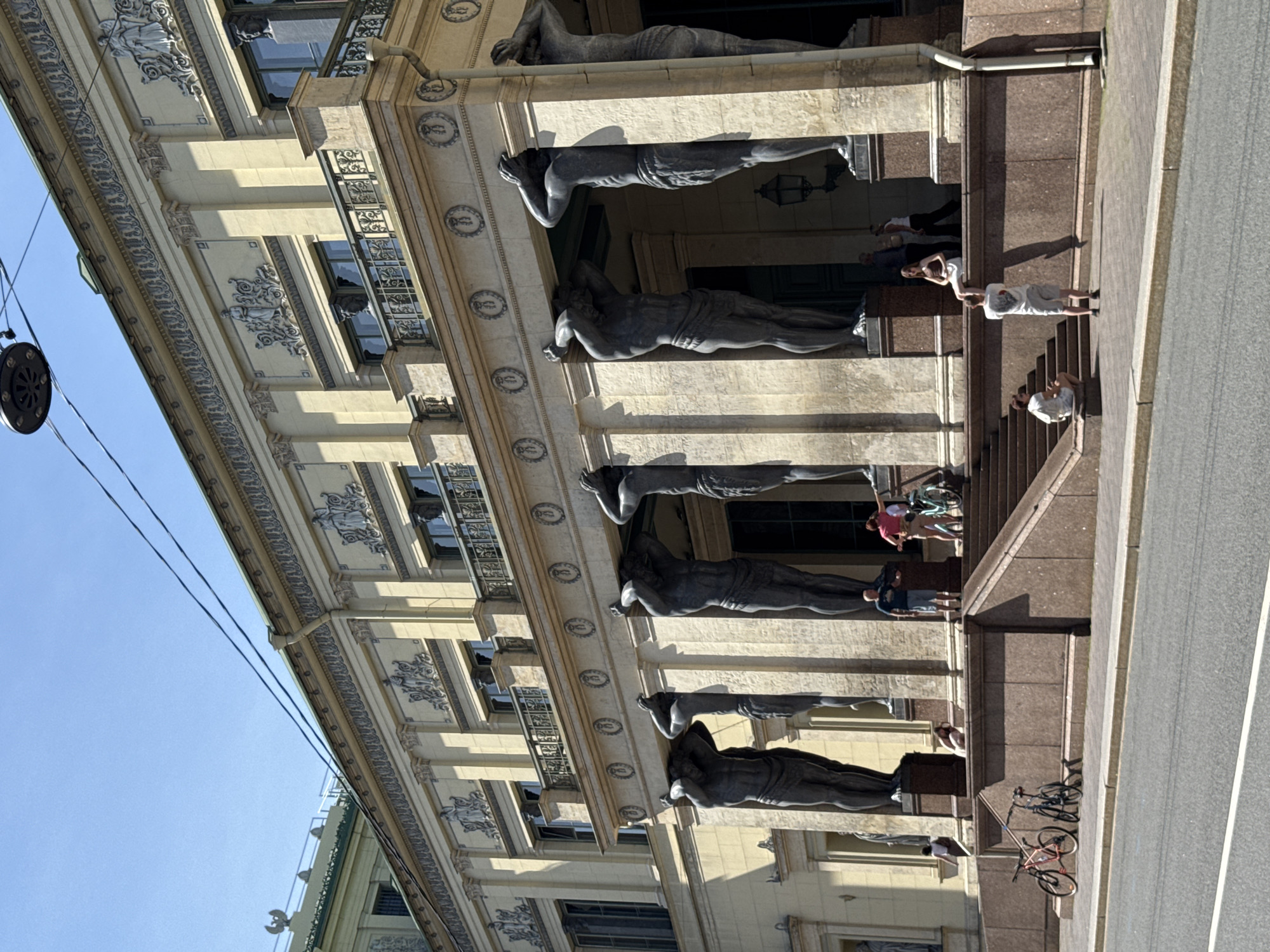 The image shows the New Hermitage building in St. Petersburg, Russia. The building's facade features classical architecture with columns and ornate decorations. In front of the building are several large, dark statues of muscular men, known as Atlantes, supporting the upper story. These statues are a prominent architectural feature.
There are several people visible on the steps and in front of the building. Some are standing and looking at the architecture, while others are interacting with each other. In the foreground, there are two bicycles parked on the sidewalk. The sky is clear and blue, indicating a sunny day. The overall scene depicts a popular tourist attraction in a historic city.