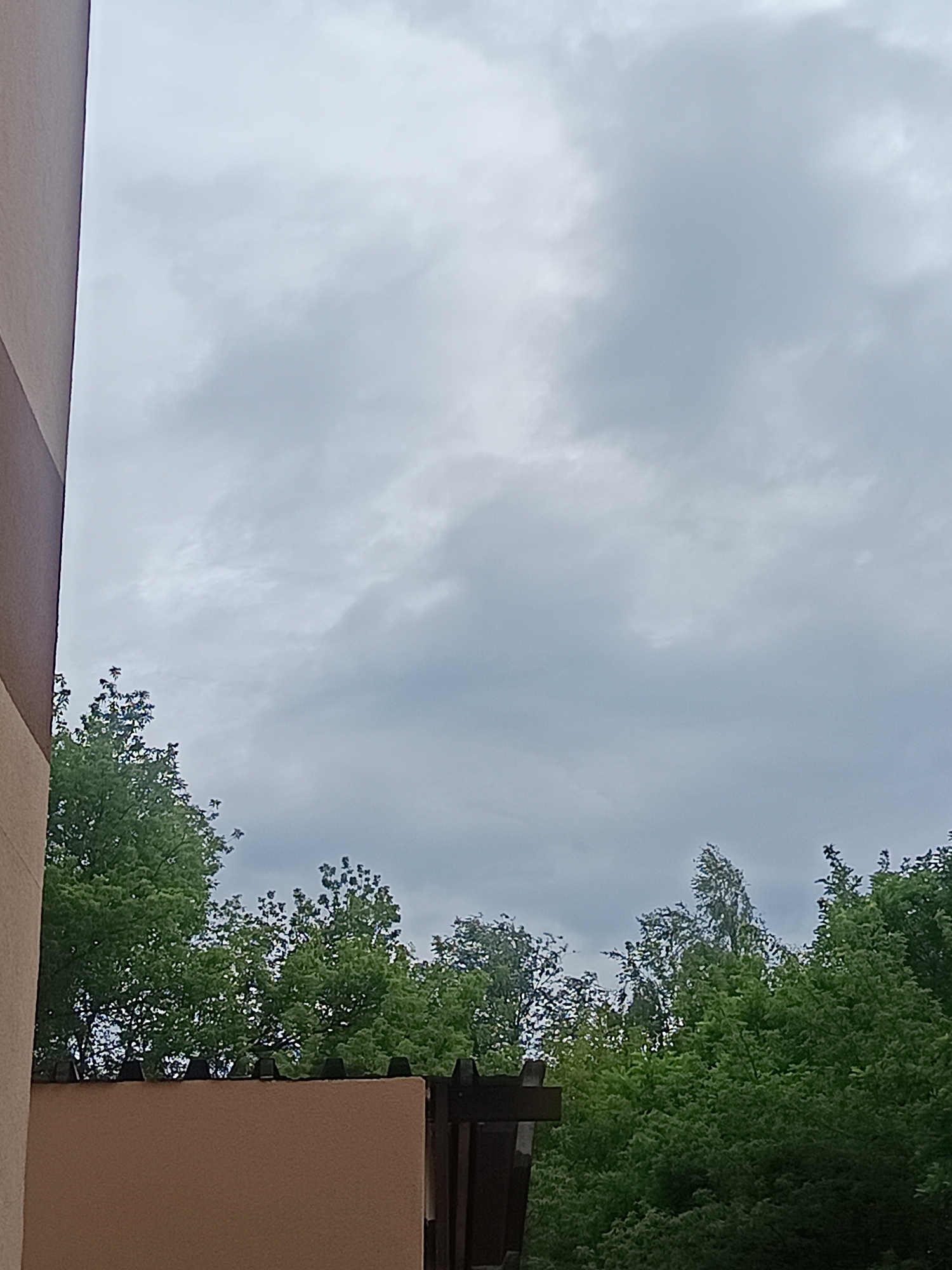 This is a close-up, vertical, eye-level shot of a wall and trees under a cloudy sky. The image appears to have been taken in Milovice, Czechia. The left side of the frame is dominated by the side of a building with a beige and brown facade. To the right of the building's facade, a low, tan-colored wall topped with black spikes is visible. Behind the wall, lush green trees fill the lower portion of the frame, extending upwards. The sky above is overcast with gray clouds, obscuring any hint of sunlight. No people are visible in the scene.