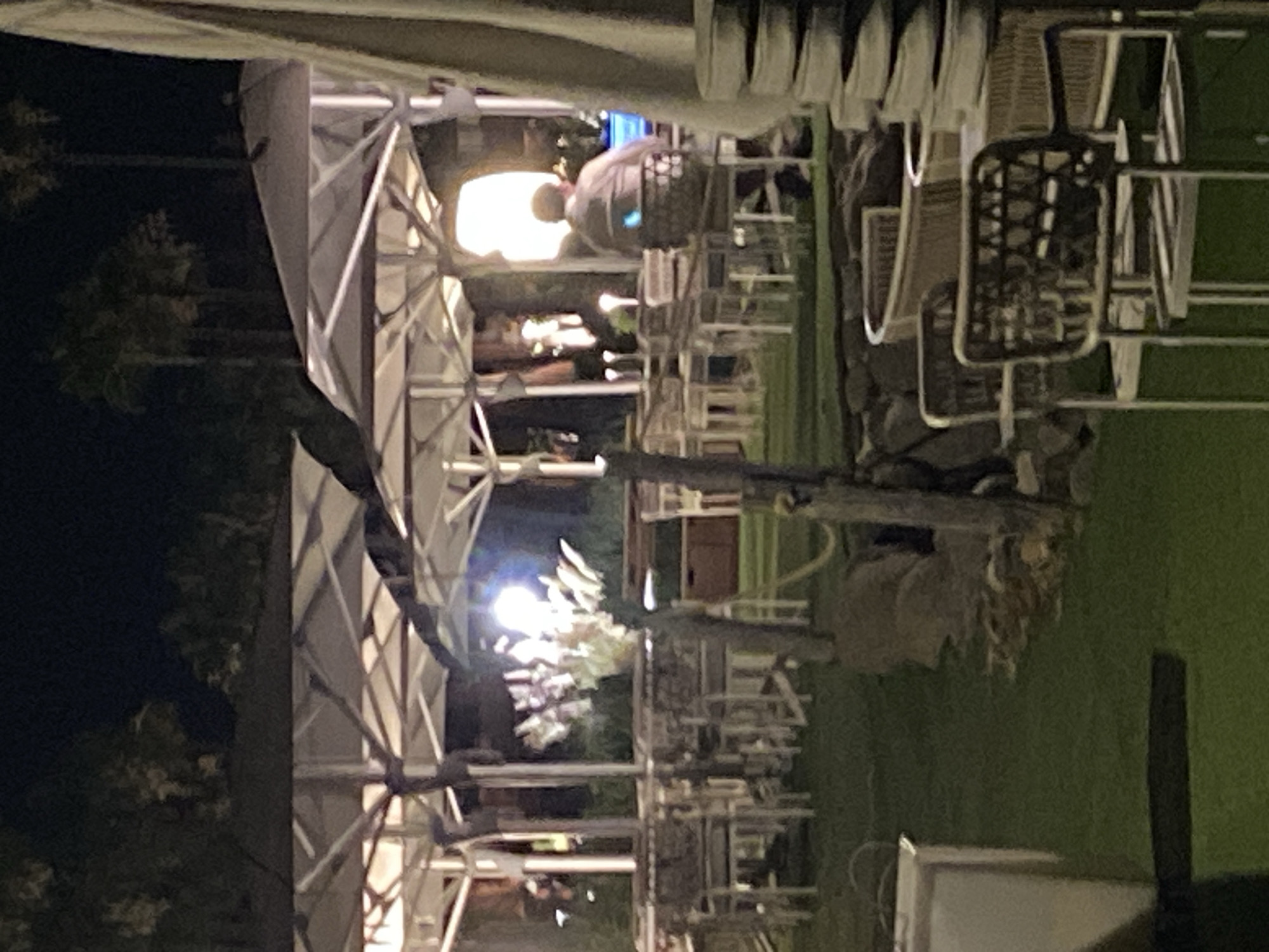 The image shows an outdoor dining area at night, possibly a restaurant or bar. There are numerous tables and chairs set up on a grassy lawn, covered by large, white umbrellas supported by metal structures. In the background, palm trees are visible against a dark sky. One person, a man wearing a light-colored shirt, is seated at a table with a laptop. The overall scene suggests a relaxed evening atmosphere, likely in a warm climate. The style of the umbrellas and the presence of palm trees are characteristic of locations along the Costa del Sol, making Estepona, Spain, a plausible setting for this scene, given its reputation for such establishments.