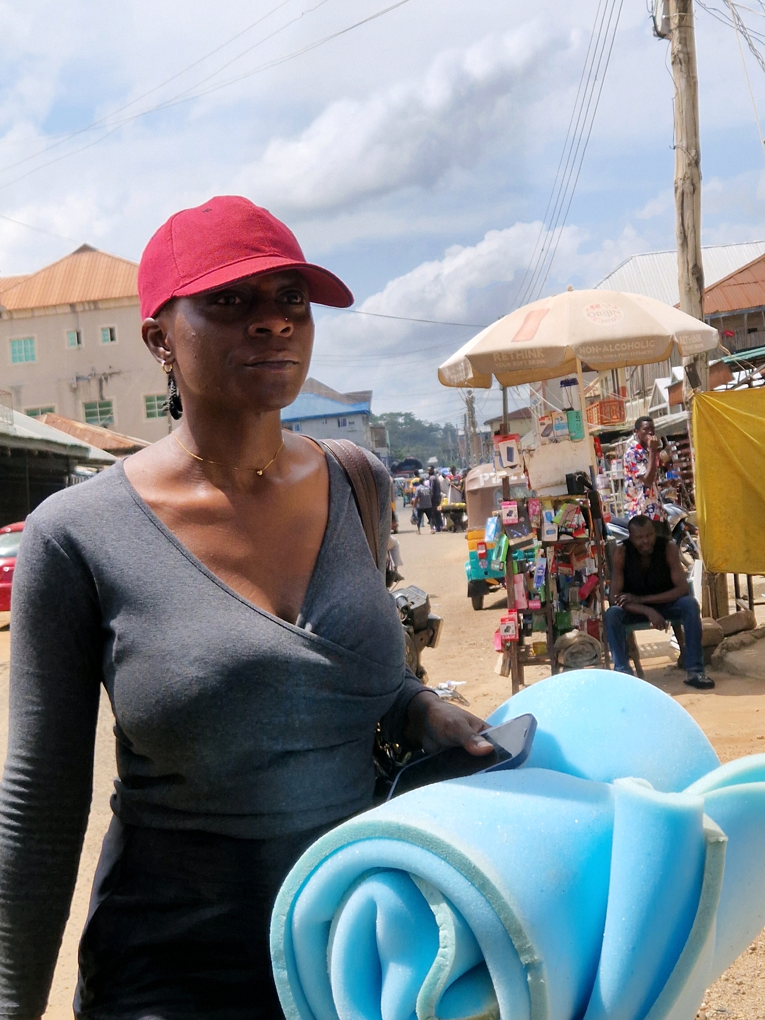 In Jos, Nigeria, a woman wearing a red baseball cap and a grey, V-neck, long-sleeved top stands outdoors, holding a rolled-up piece of light blue foam. She has on a thin gold chain necklace and hoop earrings. The woman is positioned in the foreground, with the background showing a bustling street scene. Buildings with corrugated metal roofs and windows line one side of the street, while a street vendor's stall with an umbrella displaying advertising is visible on the other. Several people are walking or standing around the stall, and a tricycle is parked nearby. A man in a dark sleeveless shirt and blue jeans sits on a stool next to the stall, looking towards the camera. The environment appears to be a commercial area with various goods displayed on shelves at the vendor's stall.