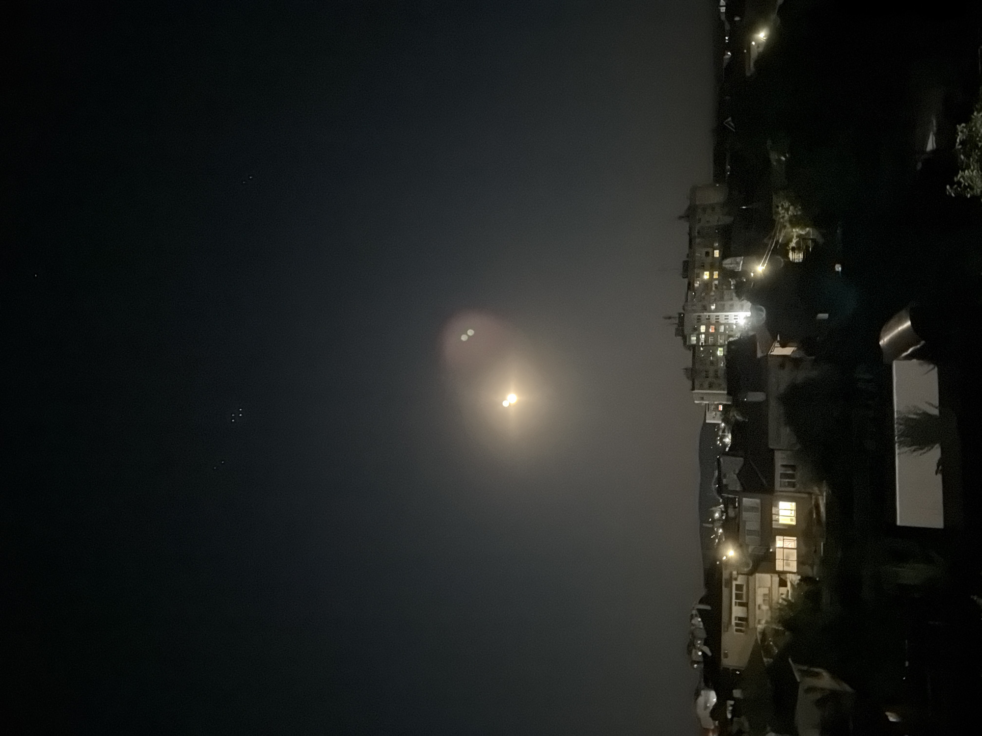 The image depicts a nighttime scene of the city of Burgas, Bulgaria. The sky is dark, with several bright stars visible in the upper left portion. A bright, diffused light source, likely the moon, is present in the upper middle of the frame, with two distinct bright spots creating a lens flare effect. Below the sky, the silhouette of a city skyline is visible, characterized by buildings of varying heights and architectural styles, some with illuminated windows. The foreground is dominated by dark shapes, likely trees and other foliage, with a partially visible white object in the lower center. The overall impression is of a tranquil urban nightscape under a starry sky.