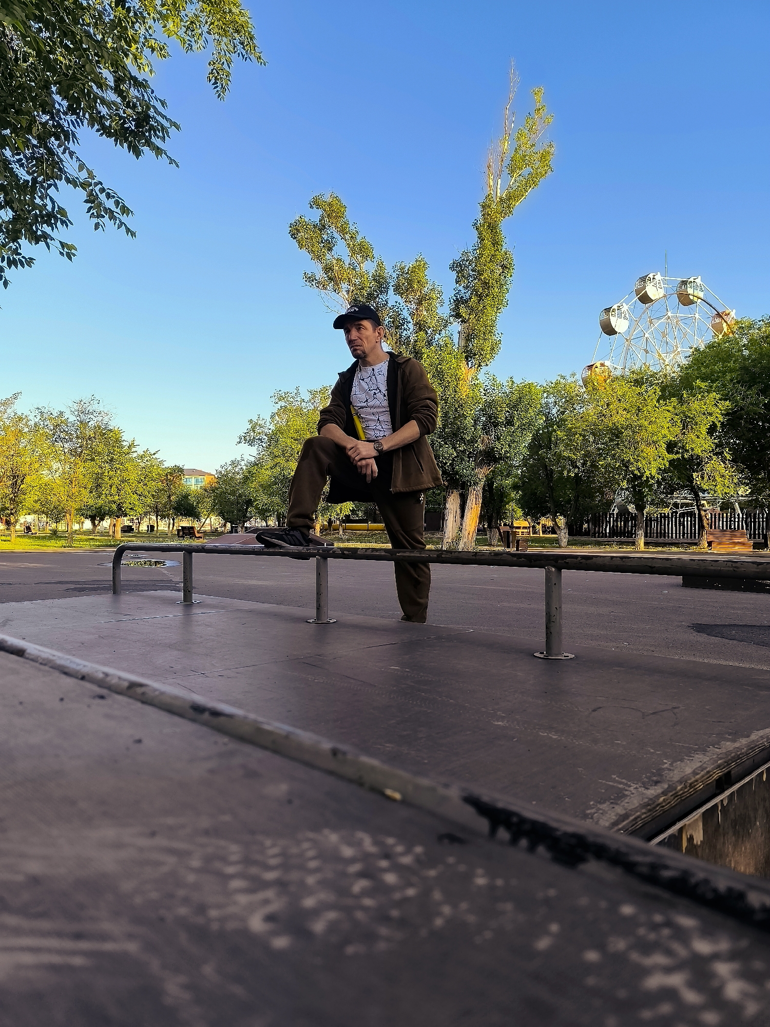 In Qarağandı city administration, Kazakhstan, a man is posed on a metal railing in a park. He is wearing a dark jacket, dark pants, a white t-shirt with a graphic print, and a black baseball cap. He is leaning with his left arm on the railing and his right arm resting on his bent knee, looking to the left of the frame. The man is positioned in the foreground, with a blurred wooden surface in front of him. Behind him, a Ferris wheel is visible in the distance, surrounded by green trees and a clear blue sky. The park appears to be a public space with pathways and other park amenities.