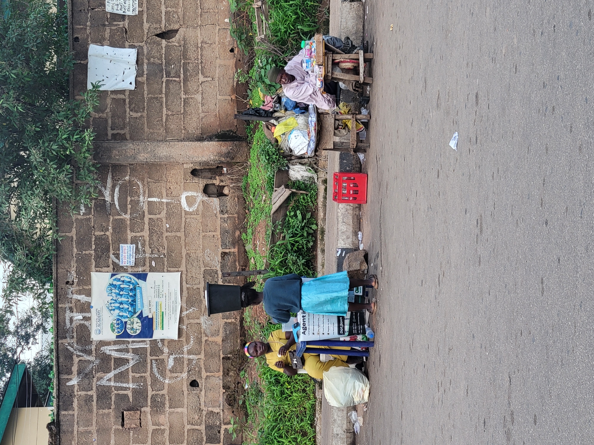 In Barakin Lerawi, Nigeria, a street scene unfolds with a weathered brick wall serving as a backdrop. Two women are engaged in commerce, one seated and wearing a yellow garment, the other standing and dressed in a blue top and skirt. The seated woman appears to be selling items from a table, while the standing woman faces away, her actions obscured. A third individual, an older man wearing a light-colored patterned shirt and a green cap, sits at another stall to the right, also appearing to be a vendor. His stall is laden with various goods, including bottled beverages and packaged items. Scattered on the ground are plastic bags, a red crate, and other debris, indicating a public thoroughfare or market area. Overgrown green vegetation flanks the wall and the vendors' setups, contrasting with the urban setting. A poster is affixed to the wall, advertising an educational institution.