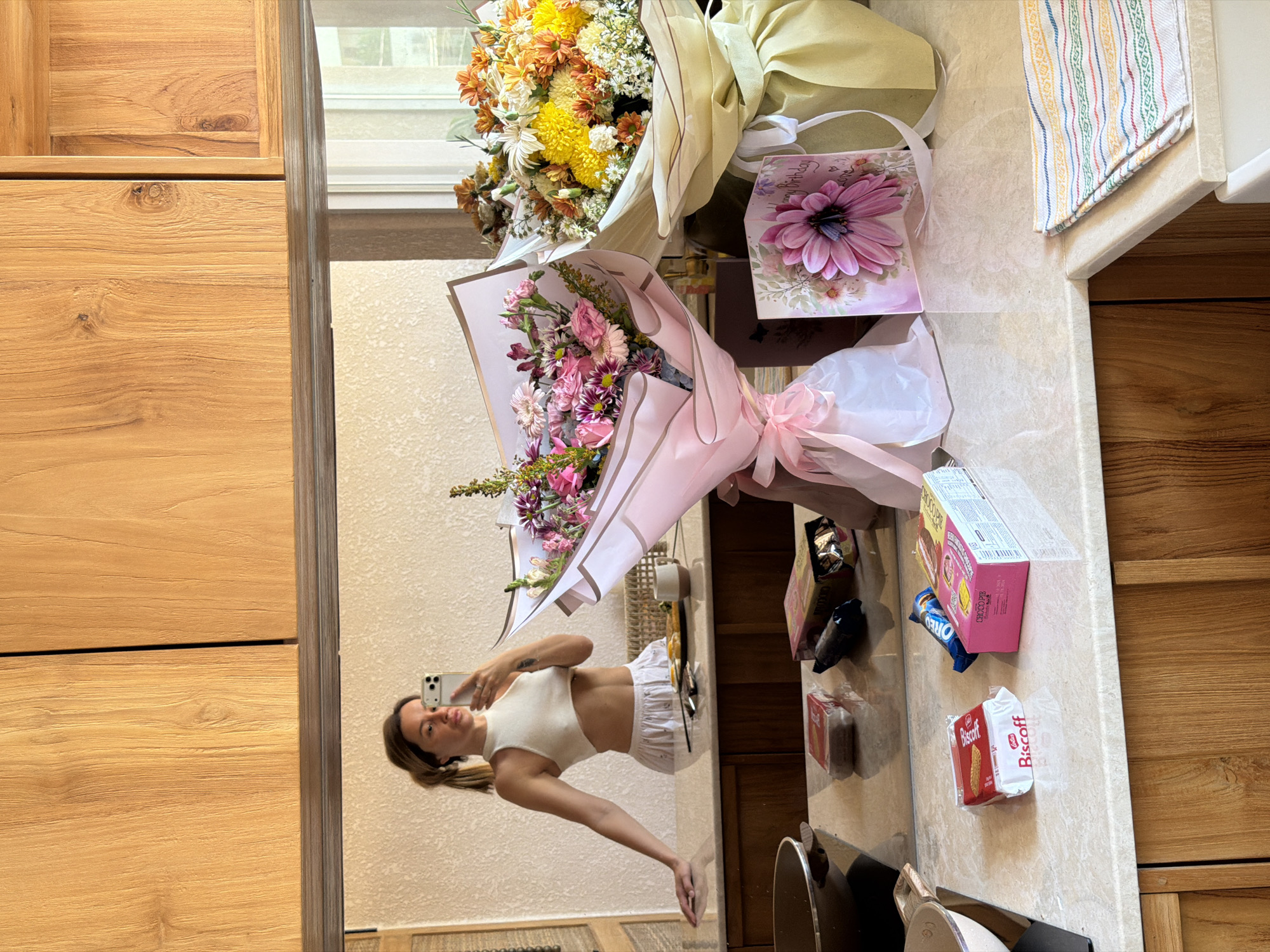 A reflective image presents a woman taking a selfie in front of a bouquet of flowers and snacks on a countertop. The countertop holds two bouquets of flowers, a birthday card, and various snacks like Choco Pie, Oreos, and Biscoff cookies. The woman is reflected in a mirror behind the countertop. Based on the furniture style, it is impossible to confirm the location as Mengwi, Indonesia, without additional evidence. The scene depicts a personal celebration, possibly a birthday.