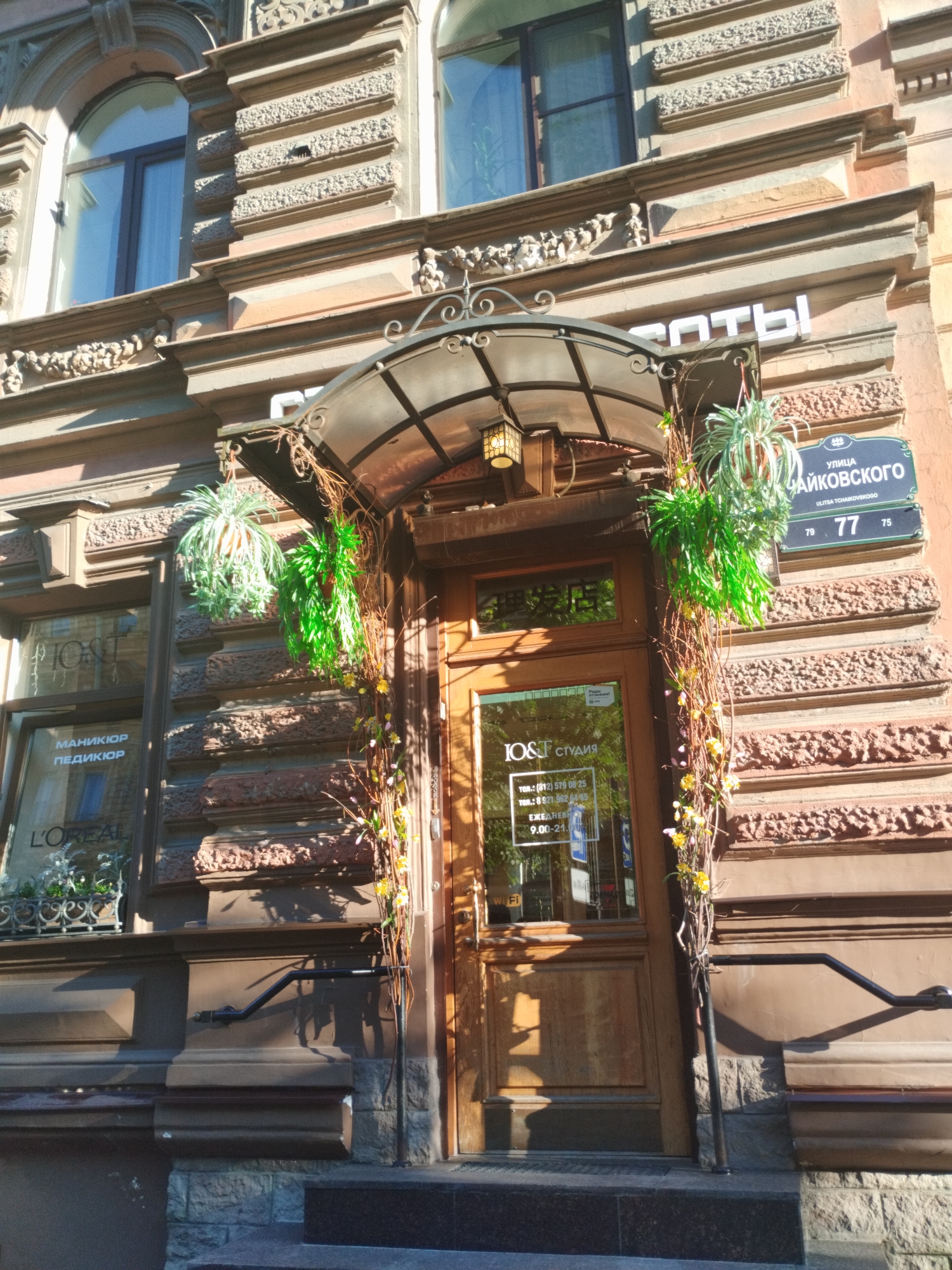 A close-up, eye-level, outdoor, natural-light photograph captures the ornate facade of a building in Saint Petersburg, Russia, likely on Tchaikovsky Street, indicated by a street sign that reads "ул. Чайковского" (ul. Chaykovskogo) with house numbers 79 and 77. The building features a rich architectural style with detailed stone carvings above the entrance and around the windows.
The focal point is a wooden double door entrance, sheltered by an arched, glass and metal awning. Hanging from the awning are two artificial plants, one a light green, feathery plant and the other a vibrant green, leafy plant. A decorative vine with small yellow flowers and branches winds its way up the side of the doorway.
To the left of the entrance, a large window displays text. Visible text includes "L'OREAL", and in Russian, "МАНИКЮР ПЕДИКЮР" (manicure pedicure), indicating the presence of a beauty salon. Another sign inside the window reads "IOST СТУДИЯ" (IOST studio) with contact phone numbers and operating hours "9.00-21.00". A small "Wi-Fi" sign is also visible on the door.
The building's facade is constructed with textured, reddish-brown bricks and stone ornamentation. Sunlight casts strong shadows on the building, highlighting the architectural details. The lower part of the image shows steps leading up to the entrance and dark-colored, stone-like materials forming the base of the building. No people are visible in the photograph.