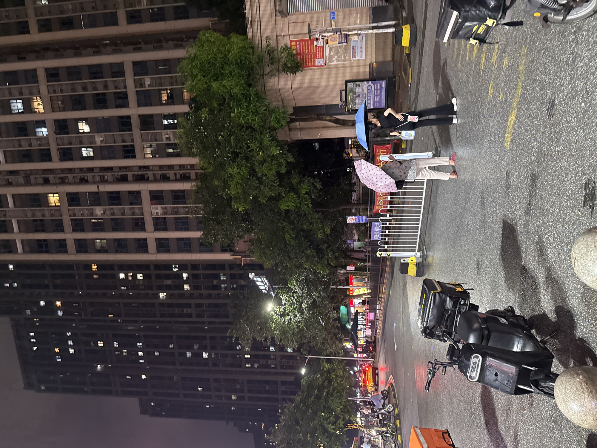 The image depicts an outdoor nighttime scene on a wet street in Haizhu District, China. Towering residential buildings with illuminated windows dominate the background, suggesting a densely populated urban area. In the foreground, a black electric scooter equipped with a large black delivery box is parked on the right side of the road. Two individuals are visible in the middle ground, standing under umbrellas on the sidewalk. One person, holding a patterned pink umbrella, is facing away from the camera. The other person, holding a blue umbrella, is facing partially away, with their right arm extended and hand on their hip. They are both wearing casual attire. A white metal barrier separates the sidewalk from the street. A yellow car is seen in the distance on the wet roadway, with its taillights illuminated. The street appears to be a commercial or mixed-use area, with some establishments or stalls visible under awnings. The overall atmosphere is that of a drizzly evening in a Chinese city.