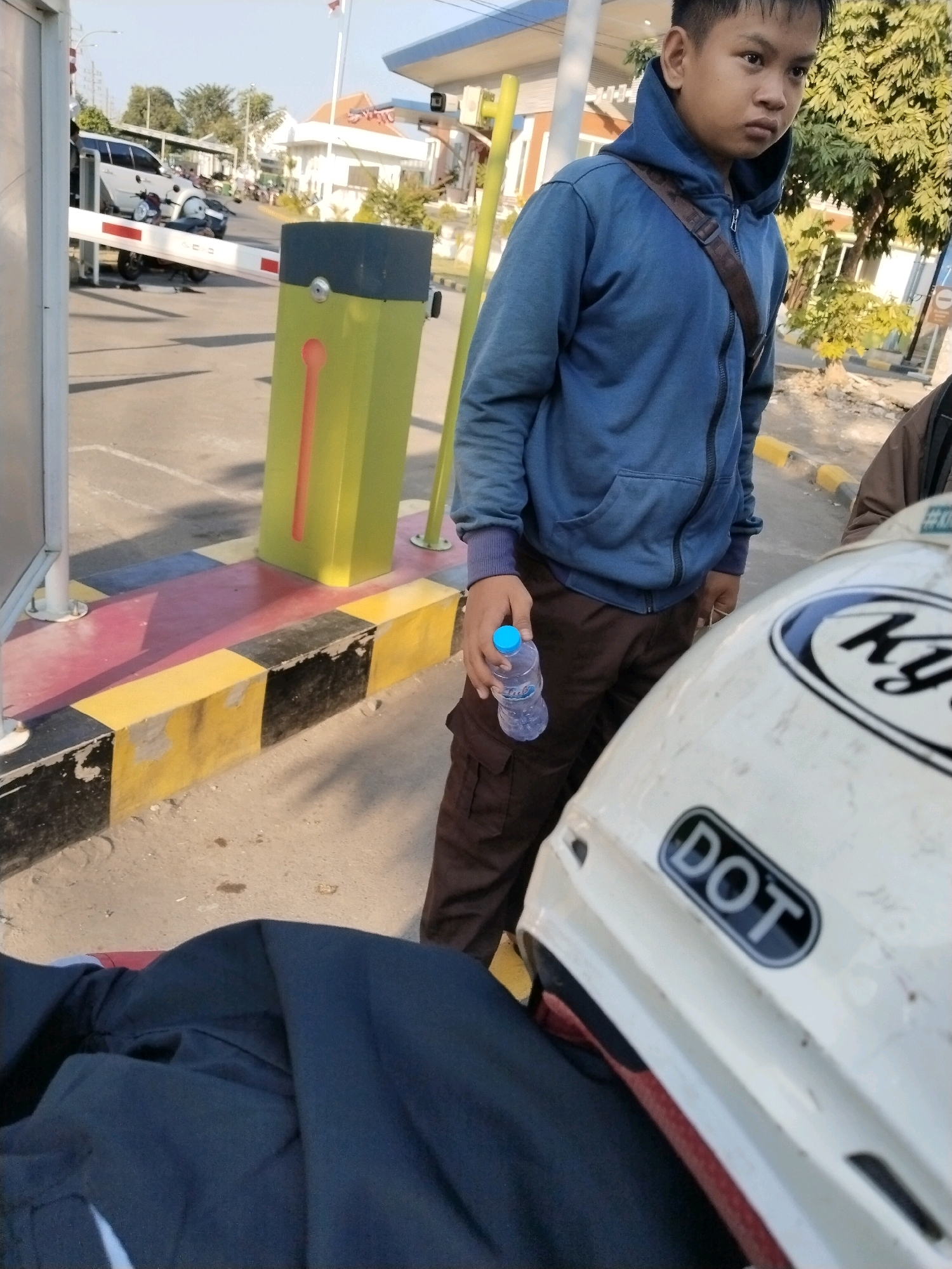 In Wonocolo, Indonesia, a young male in a blue hooded jacket and brown cargo pants stands outdoors, holding a plastic water bottle. He is positioned near a yellow and gray automated gate. In the foreground, a white motorcycle helmet with a "DOT" sticker is visible. In the background, a white SUV and other motorcycles are present, along with buildings and trees under a clear sky.