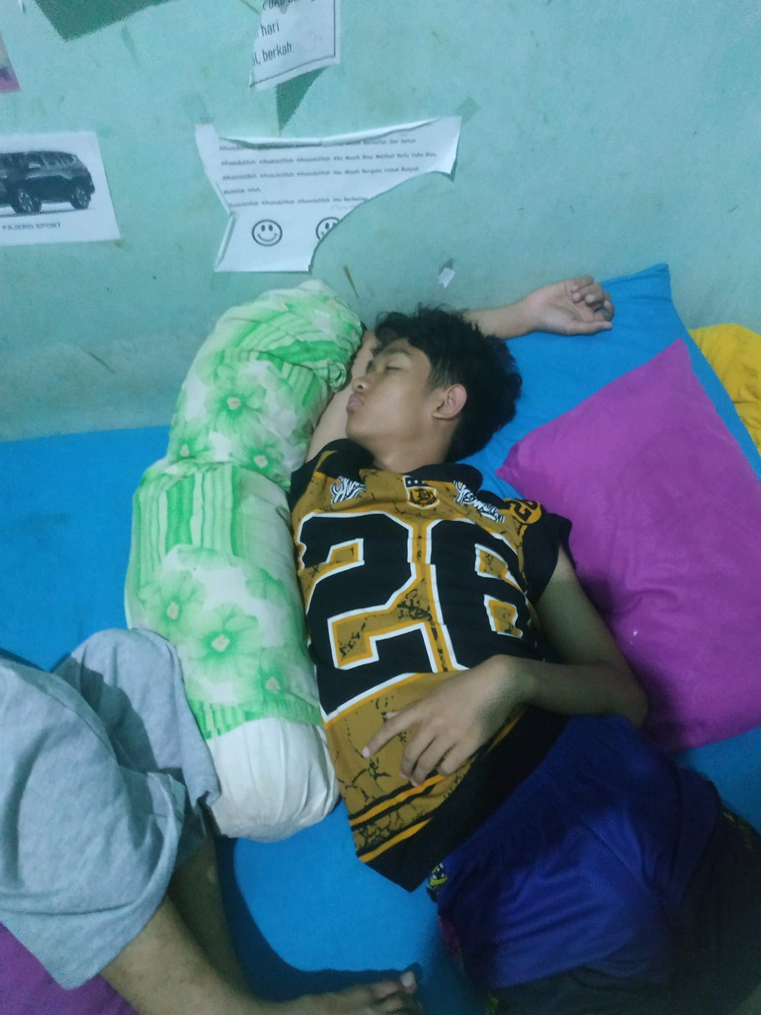 The image shows a young person sleeping on a bed, likely in a room in Pacul, Indonesia. The person is lying on their side, facing away from the camera, with their eyes closed and their mouth slightly open. They are wearing a black t-shirt with a gold and white graphic design, and blue shorts. A large green and white floral pillow is positioned behind their head, and a blue pillow is under their head. To the right of the person, a purple pillow is visible. In the background, a light blue wall is adorned with several pieces of paper, including a picture of a black SUV labeled "PAJERO SPORT," a piece of paper with some Arabic writing, and a piece of paper with smiley faces and more Arabic writing. The floor appears to be covered by a light blue sheet or mat, with a purple sheet visible at the bottom edge of the frame. The overall impression is of a casual, domestic setting.