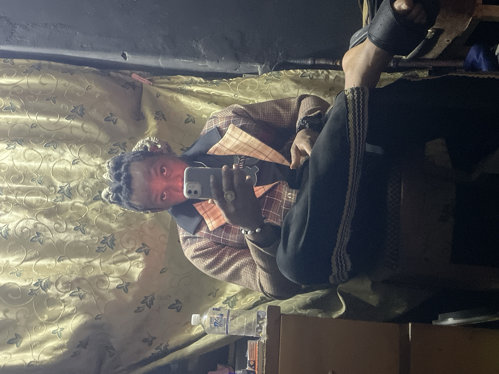 The image shows a man taking a selfie in a room. He is wearing a plaid blazer and black pants with a white stripe. He is also wearing a ring and a bracelet. He is holding a cell phone in his left hand. His feet are visible in the upper right corner, with one foot clad in a black sandal. The room is dimly lit and has patterned gold and blue wallpaper. A water bottle and other personal items are visible on a shelf near the bottom of the frame. The man appears to be in a confined space with limited headroom, judging by the proximity of the ceiling. The exact location in Jos, Nigeria cannot be determined from the image.