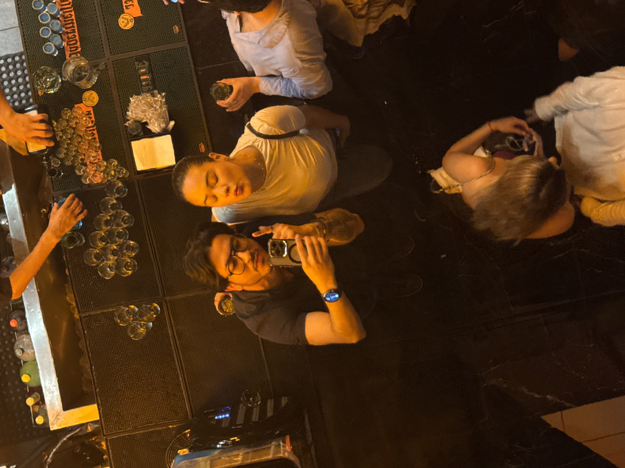 The image, likely taken in Puebla, Mexico, shows an overhead view of a bar scene. A bartender is preparing drinks behind a bar with bottles and glassware. Three people, a man and two women are together, with the man facing the camera and taking a photo with his phone. He wears a black shirt and eyeglasses and has a watch on his left wrist. One of the women stands next to him and wears a white t-shirt. The other two people, also likely women, are on the right side of the frame. The woman on the left is on her knees, looking at the floor with a phone in her hand. The last woman is behind the woman on her knees and is wearing a white shirt. The bar area features a textured mat surface where the glasses and drinks are placed.